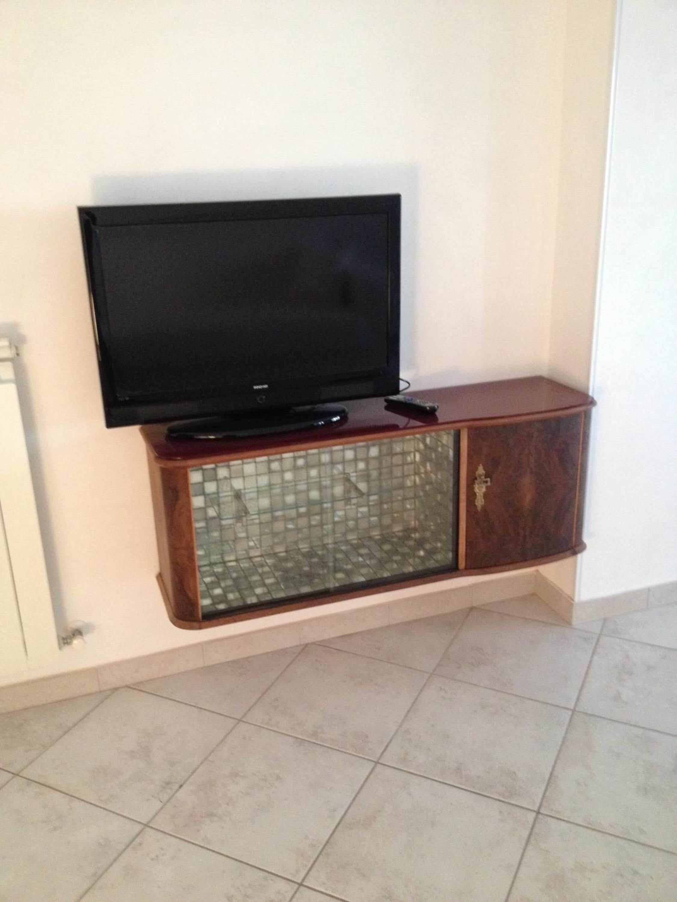 Un televisor de pantalla plana montado en un mueble de madera con puerta de cristal, ubicado frente a una pared blanca en una habitación con suelo de baldosas.