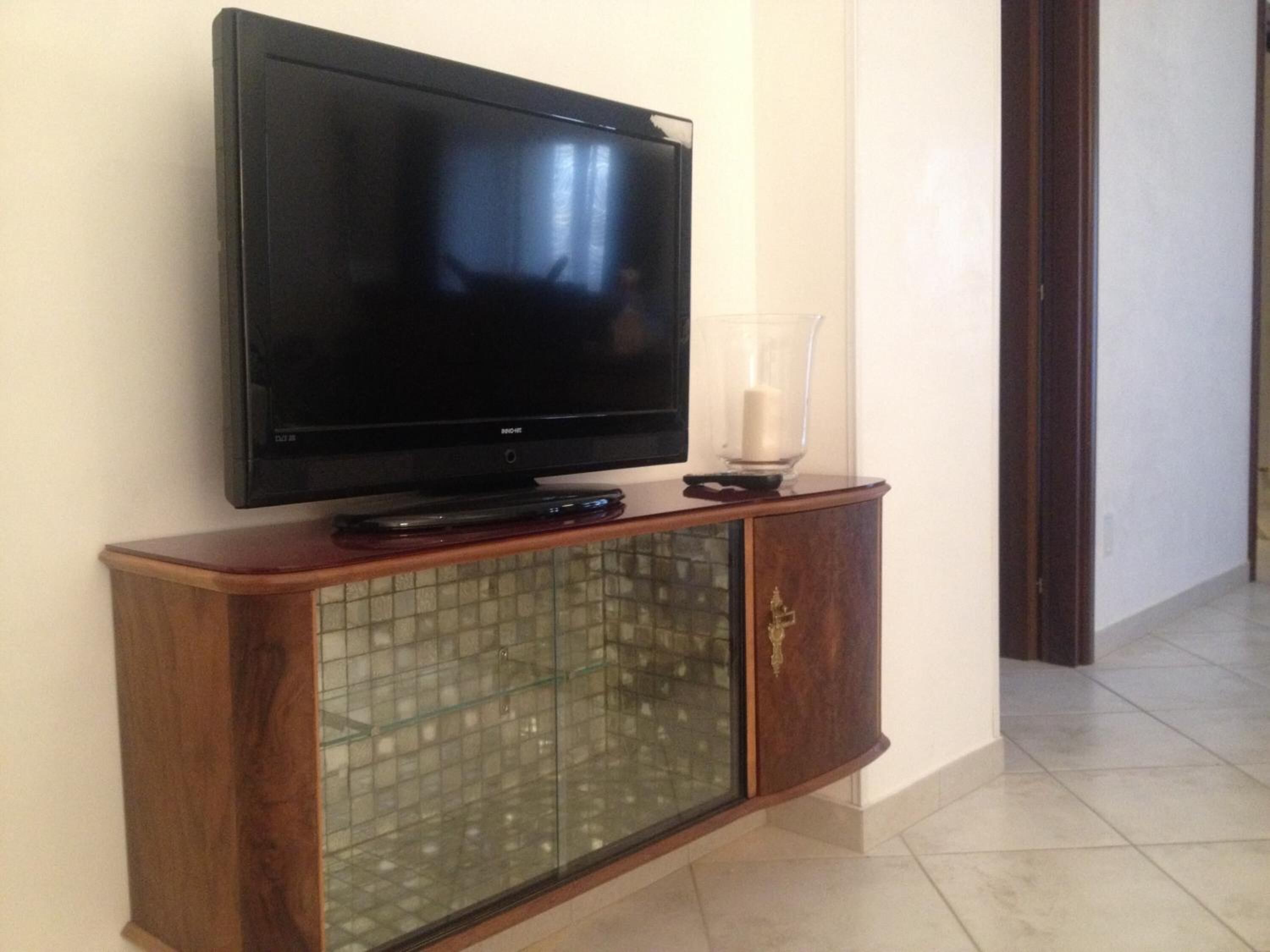 Un televisor de pantalla plana sobre un mueble de madera con puerta de vidrio, ubicado en un pasillo con piso de baldosas.