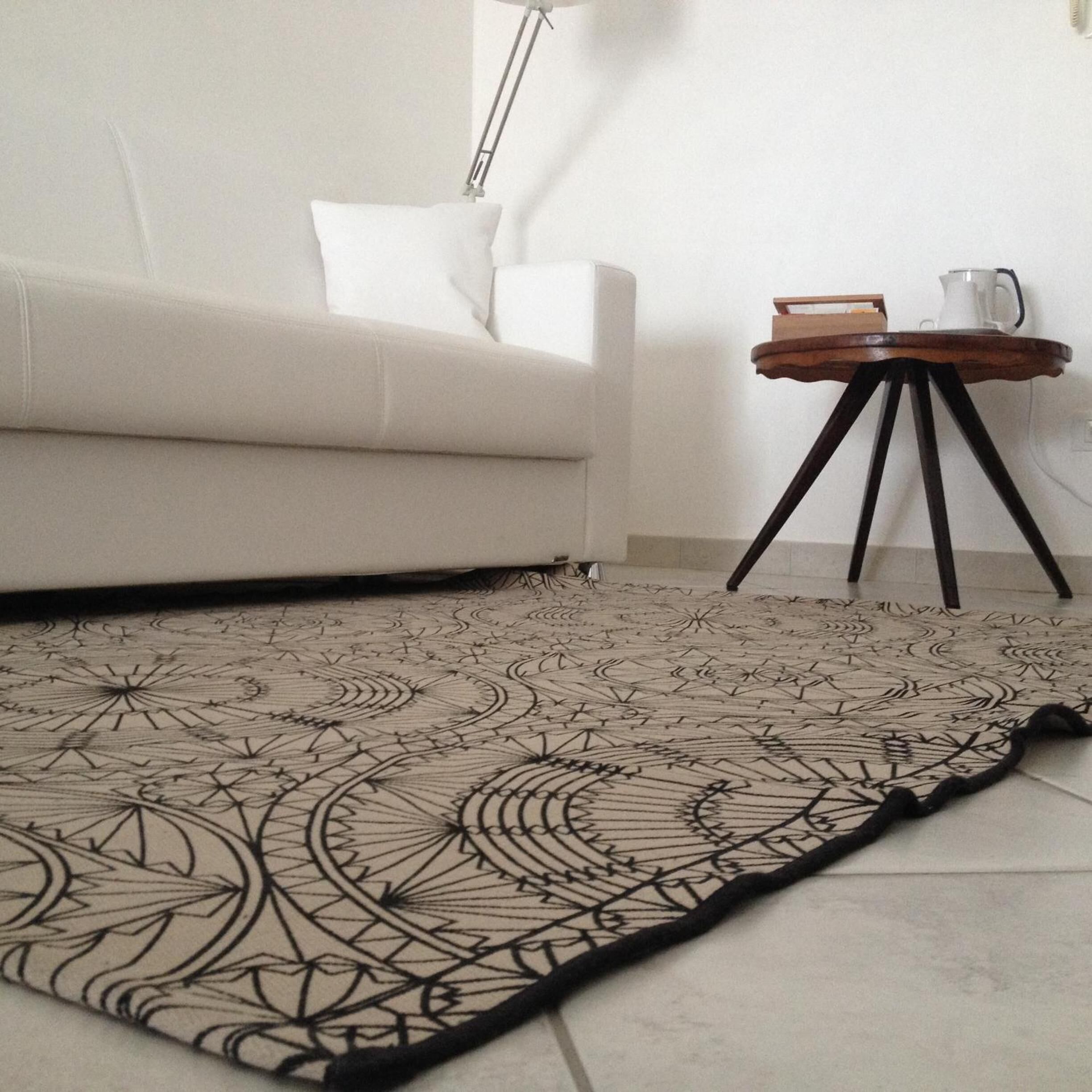 Un salon avec un canapé blanc, un tapis à motifs, une petite table d’appoint en bois sur laquelle se trouve une bouilloire, et une lampe.
