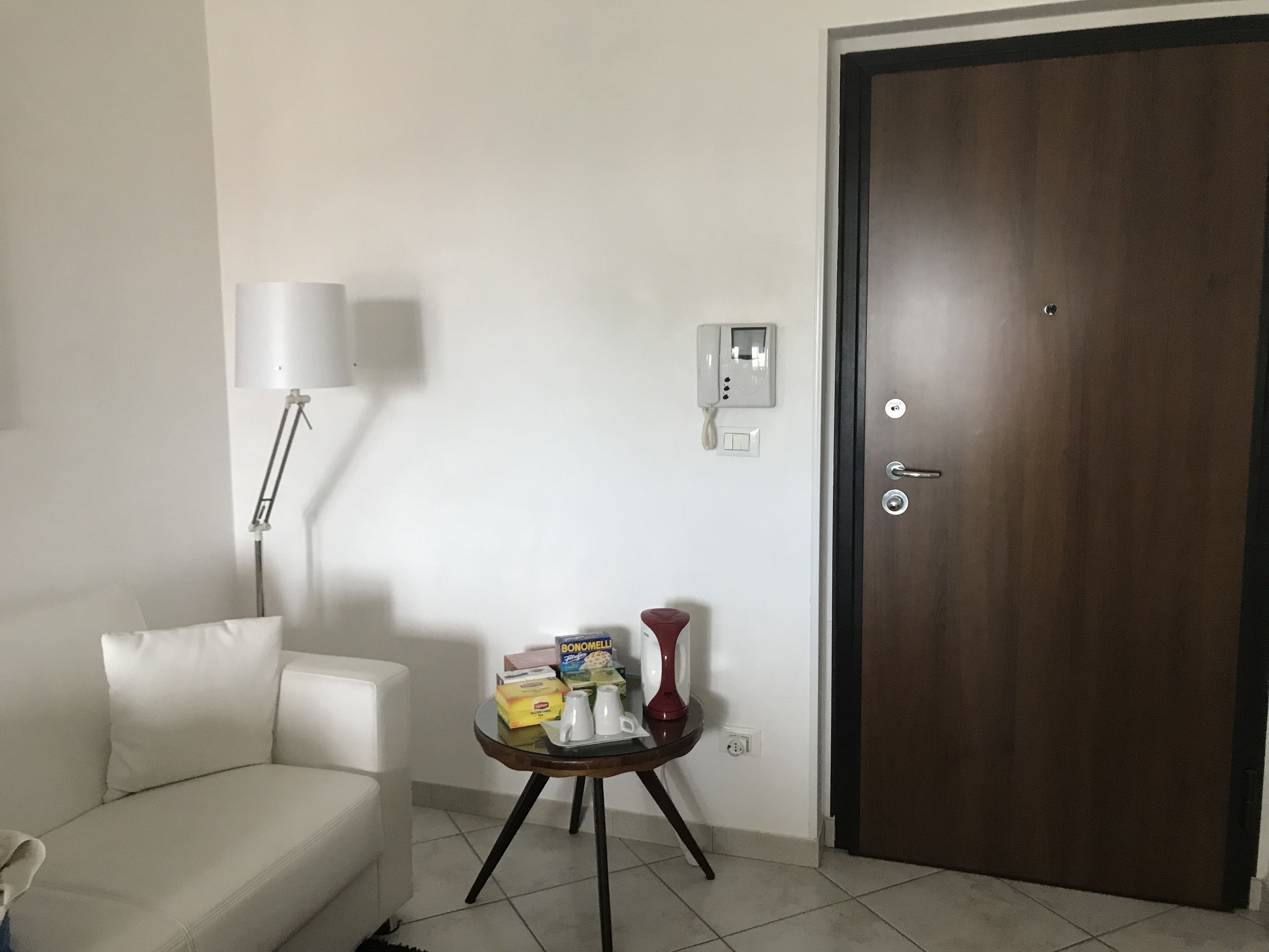 Une pièce avec un canapé blanc, une petite table ronde sur laquelle se trouvent divers objets, une lampe, un téléphone fixé au mur et une porte en bois.