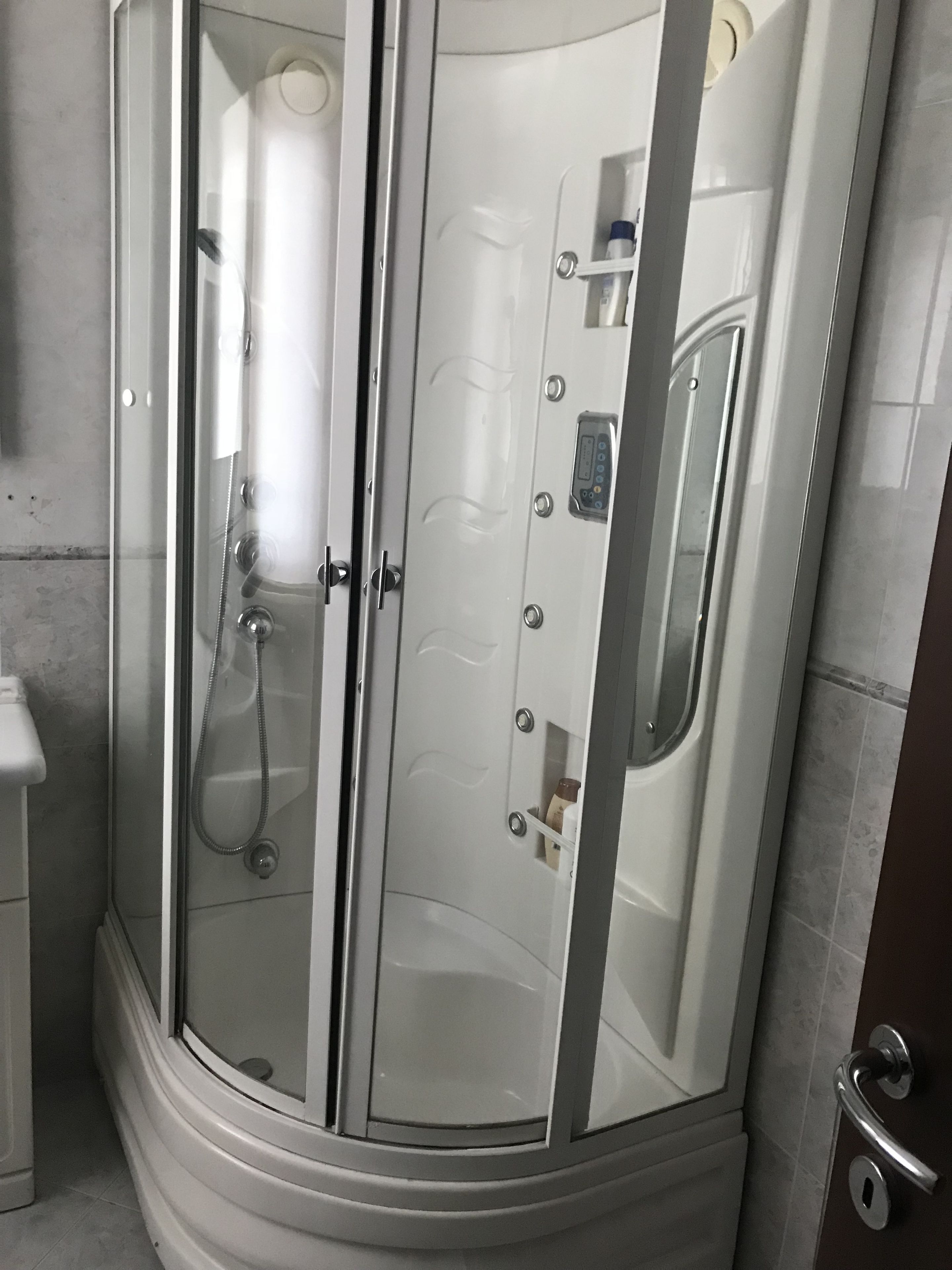 A glass-enclosed shower stall with a handheld showerhead, a control panel, and a towel rack.