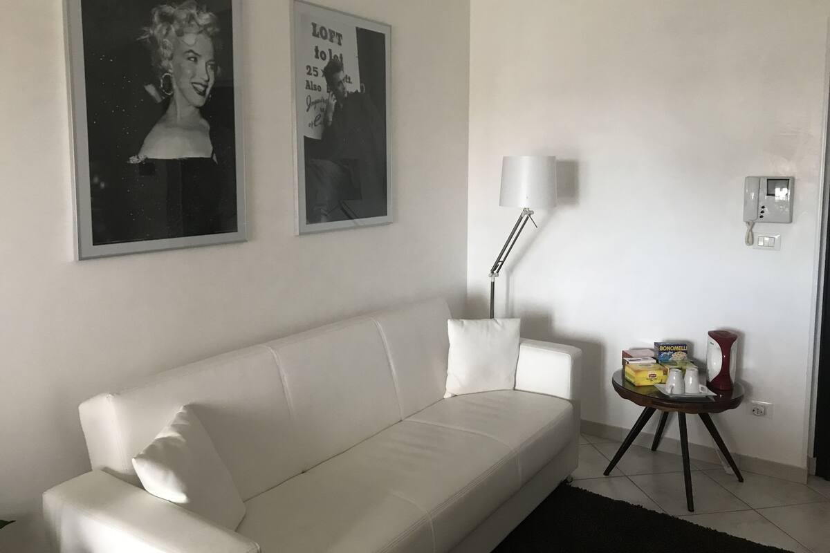 A white sofa in a living room with two framed pictures on the wall, a floor lamp, a small table with a red lamp, and a wall-mounted phone.