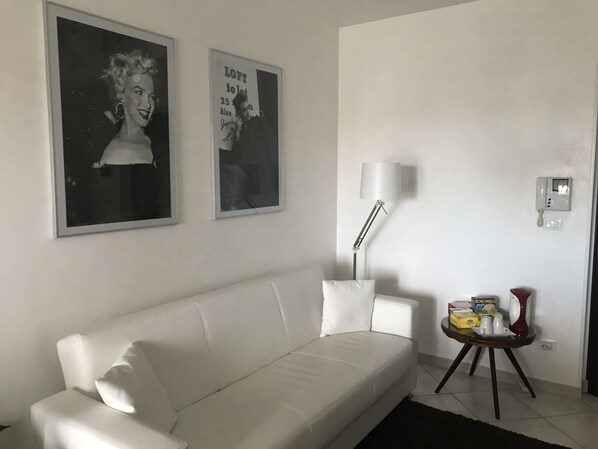 A white sofa in a living room with two framed pictures on the wall, a floor lamp, a small table with a red lamp, and a wall-mounted phone.