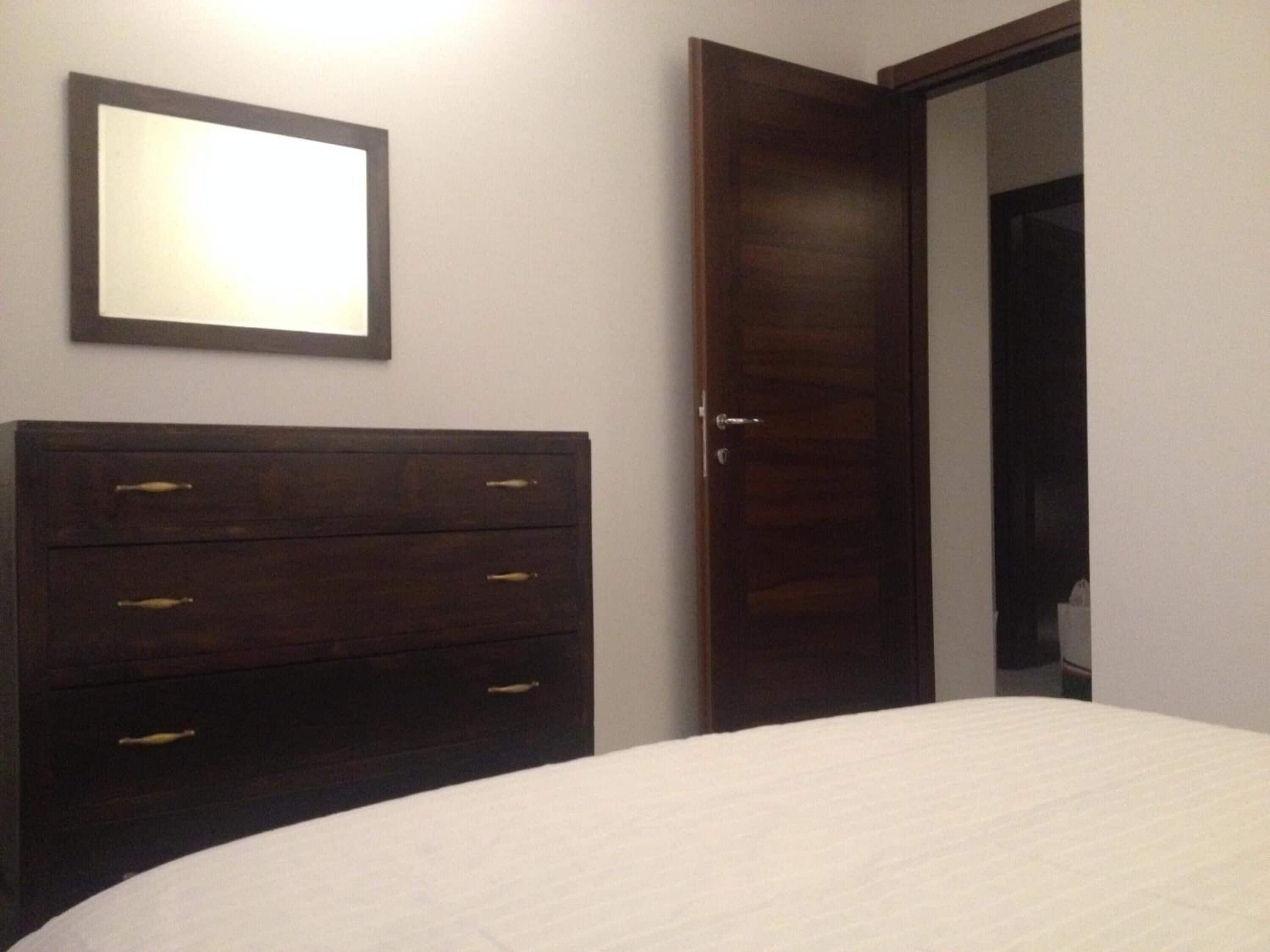 Een slaapkamer met een donkere houten dressoir, een wit bed en een houten deur.