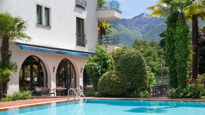 A swimming pool with a wooden deck, outdoor seating, and a view of mountains. - Aparthotel al Lago (Locarno)
