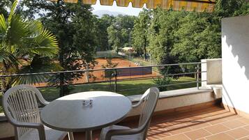 En balkon med bord og stole, udsigt over en tennisbane og frodig vegetation.