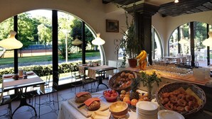 A breakfast buffet with various fruits, pastries, and bread, set in a room with large windows and a view of the outdoors. - Aparthotel al Lago (Locarno)