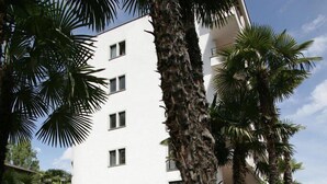 A white multi-story building with balconies, surrounded by lush greenery and pink flowering bushes. - Aparthotel al Lago (Locarno)