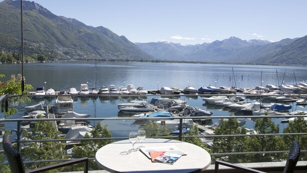 A terrace with a table, a magazine, and a glass of water. The terrace overlooks a marina with boats and mountains in the background. - Aparthotel al Lago (Locarno)