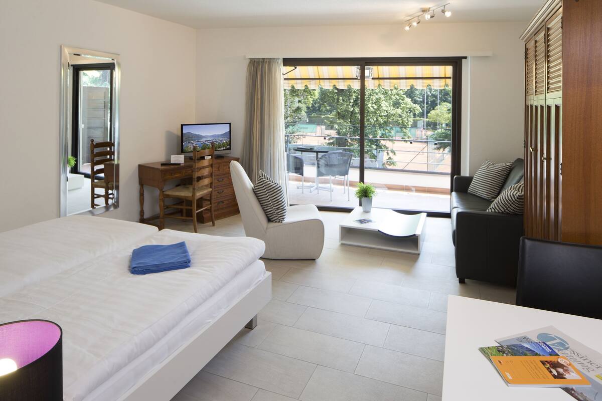 A modern hotel room with a bed, a desk, a chair, a sofa, and a balcony with a view.