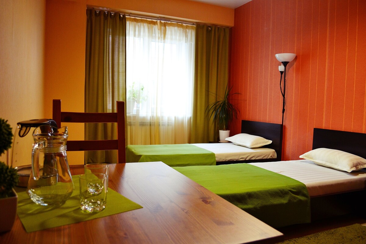 Habitación de hotel con dos camas, una mesa de madera, una jofaina y un vaso sobre un tapete verde.