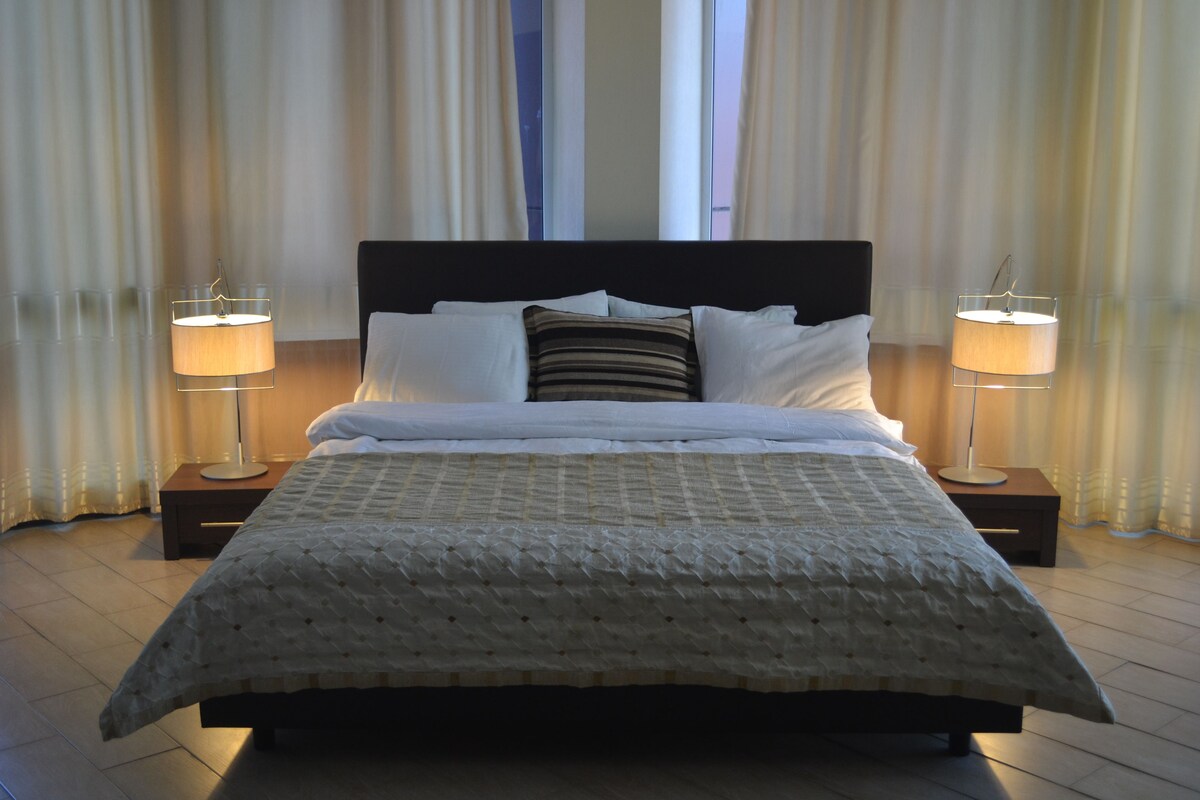 Una cama bien hecha con edredón blanco y almohadas, flanqueada por dos lámparas de noche sobre mesitas de madera.