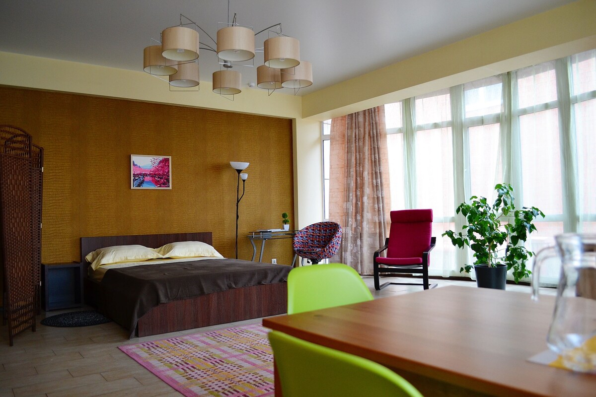 Habitación de hotel con cama, escritorio, silla, lámpara, planta y ventanas grandes.