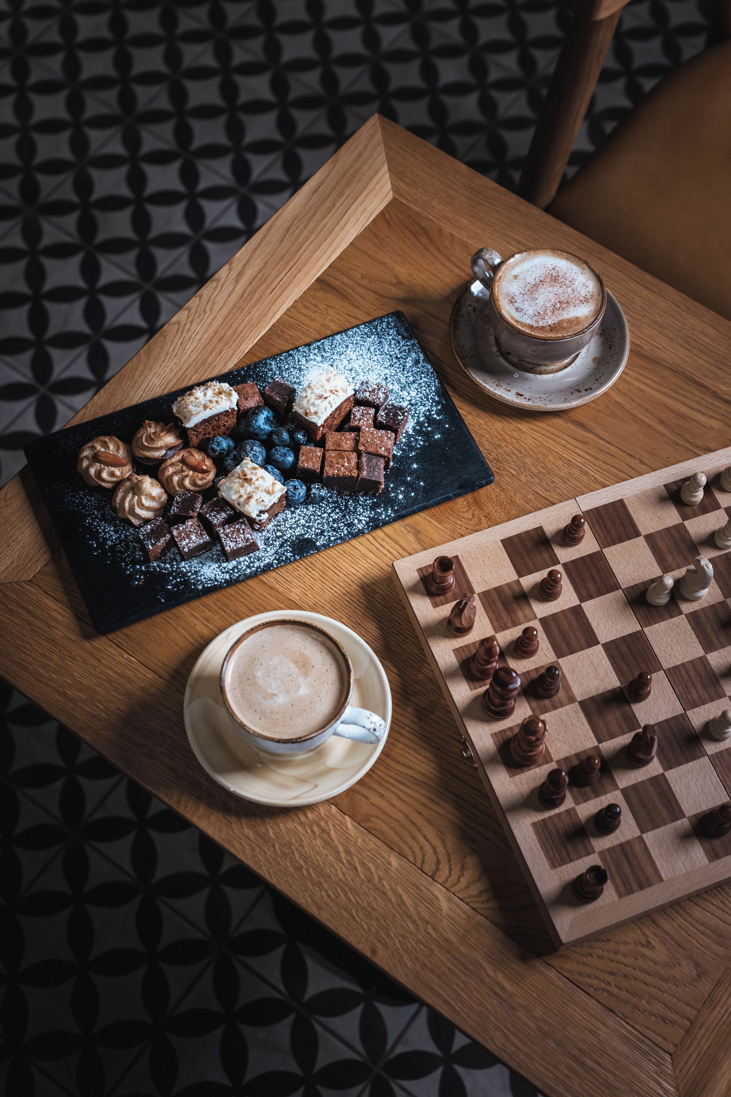A wooden table with a chessboard, a cup of coffee, and a dessert tray.