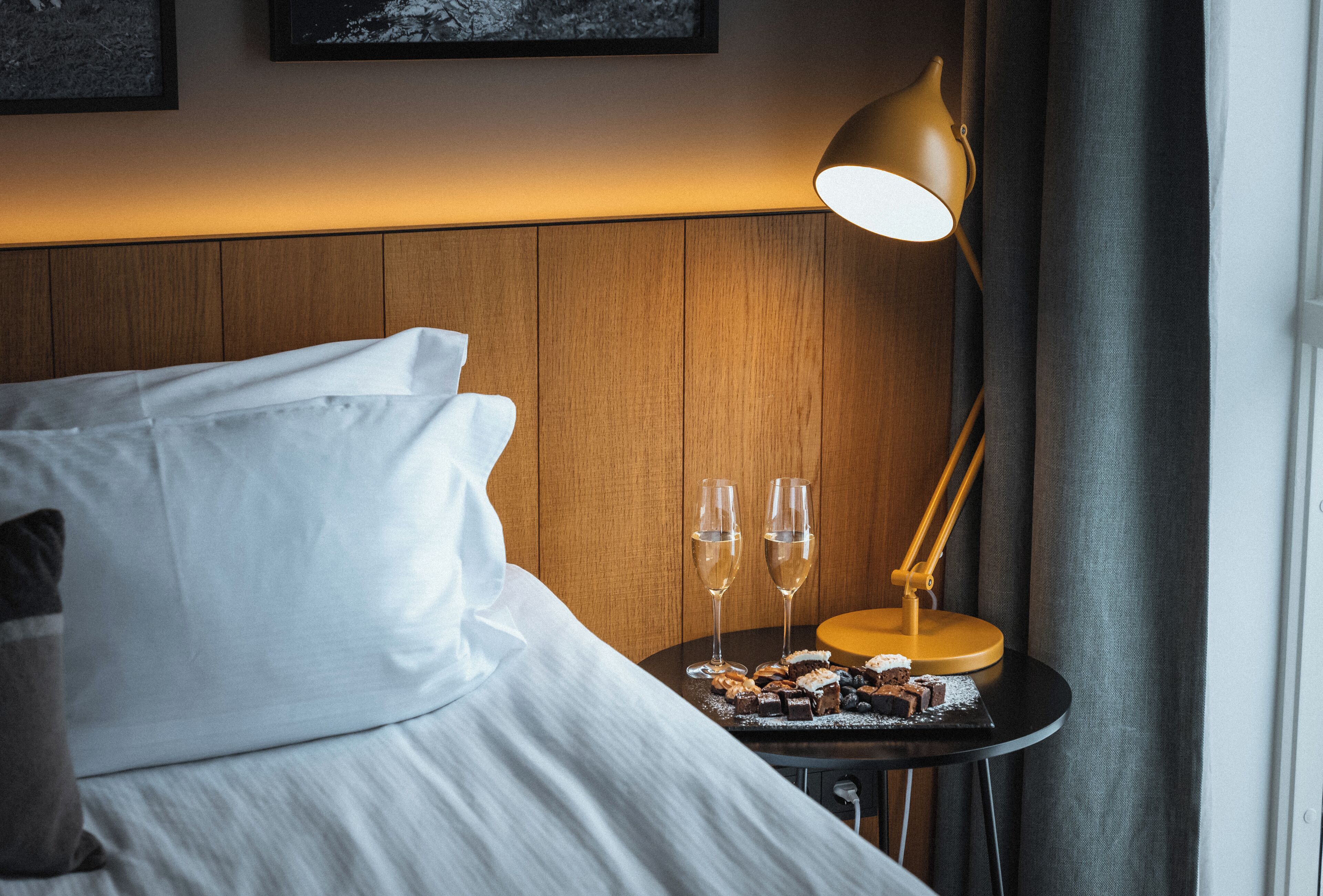 A neatly made bed with white linens, a small table with a lamp and two glasses of water, and a tray with desserts.