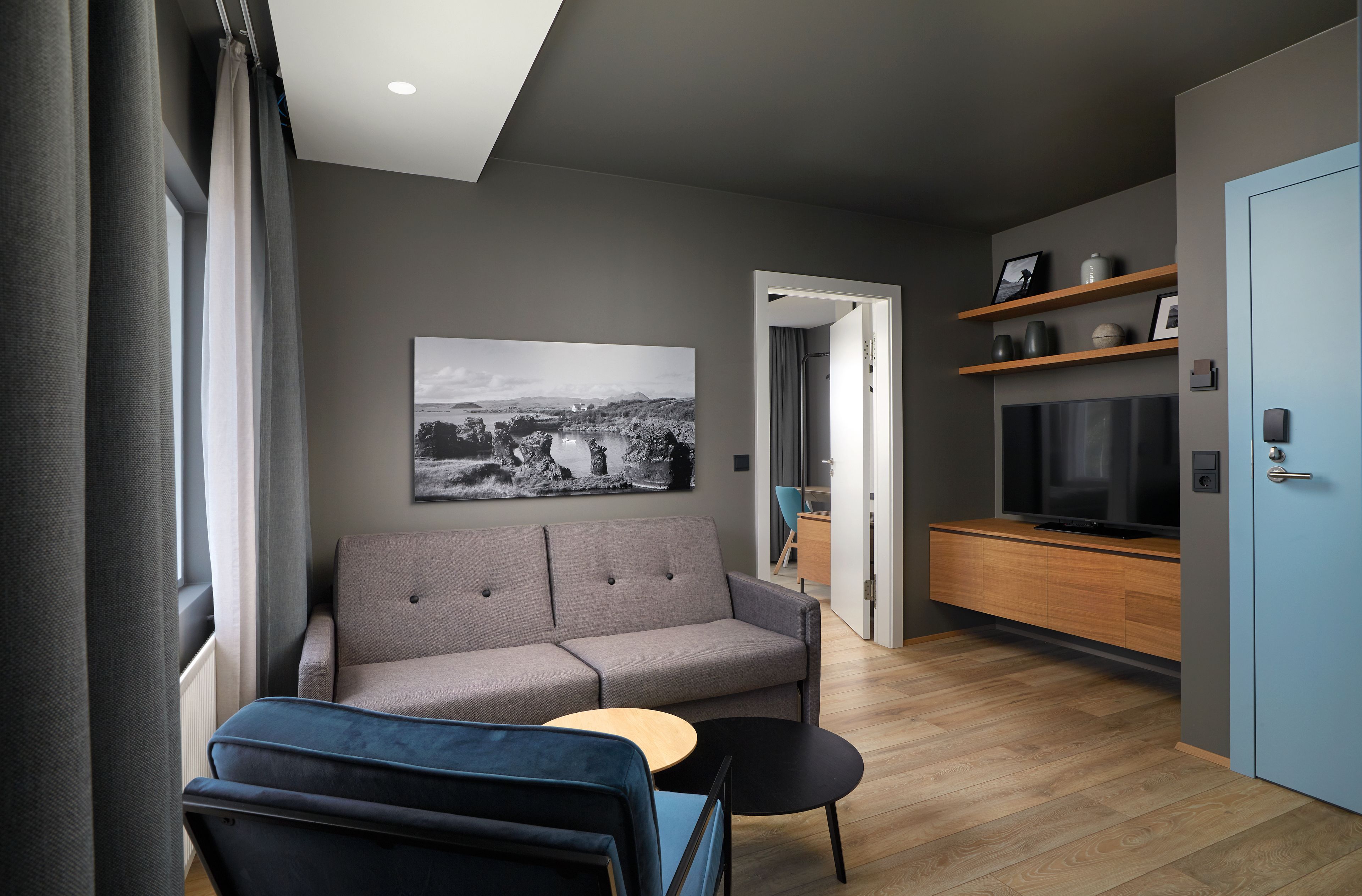 Una sala de estar moderna con un sofá gris, un sillón azul y una pequeña mesa de madera. Hay una fotografía grande en blanco y negro en la pared, un televisor sobre un mueble de madera y una puerta azul.