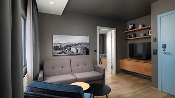 Una sala de estar moderna con un sofá gris, un sillón azul y una pequeña mesa de madera. Hay una fotografía grande en blanco y negro en la pared, un televisor sobre un mueble de madera y una puerta azul.