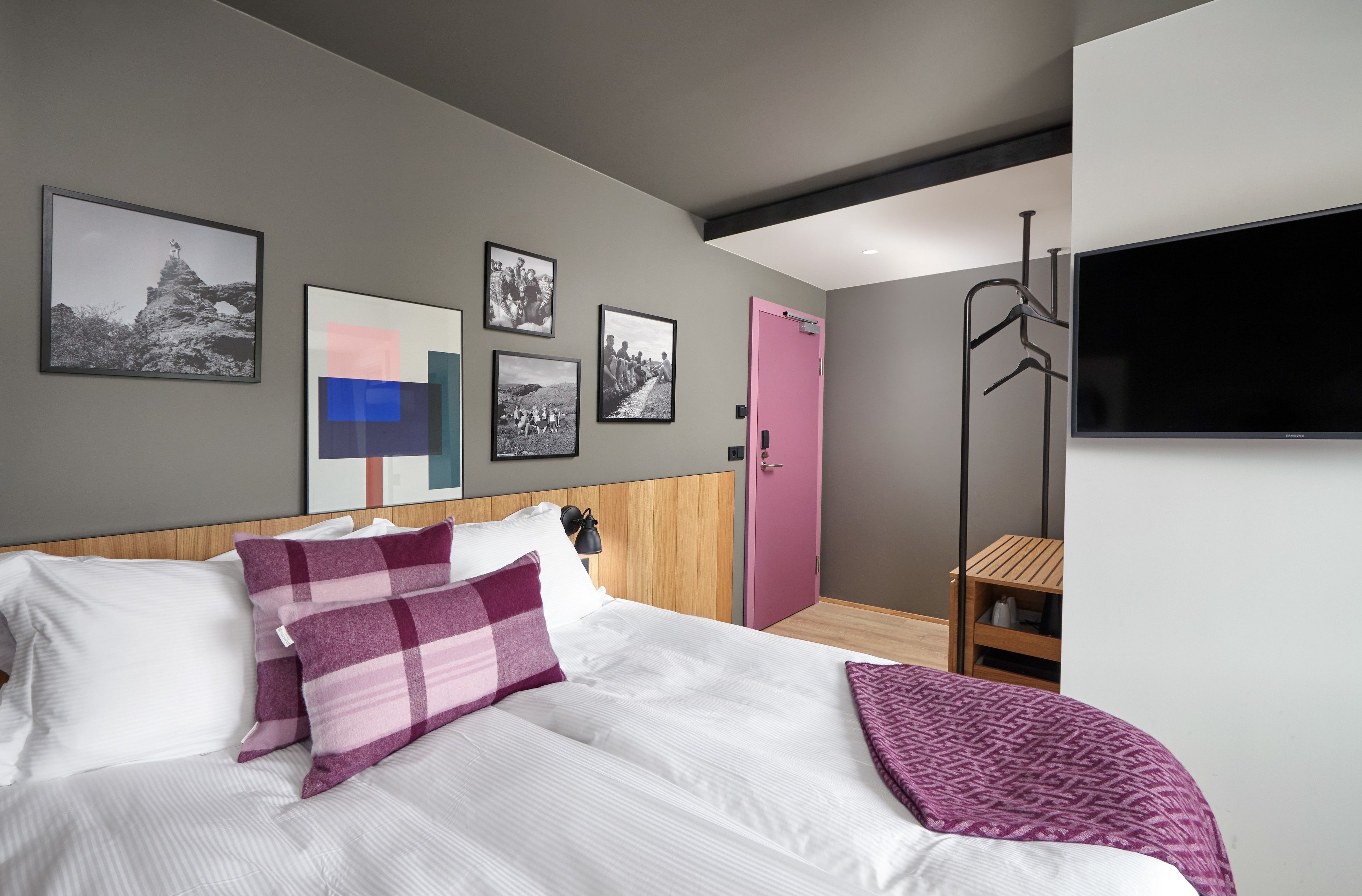 A modern hotel room with a large bed, a wooden headboard, a pink door, and a flat-screen TV mounted on the wall.