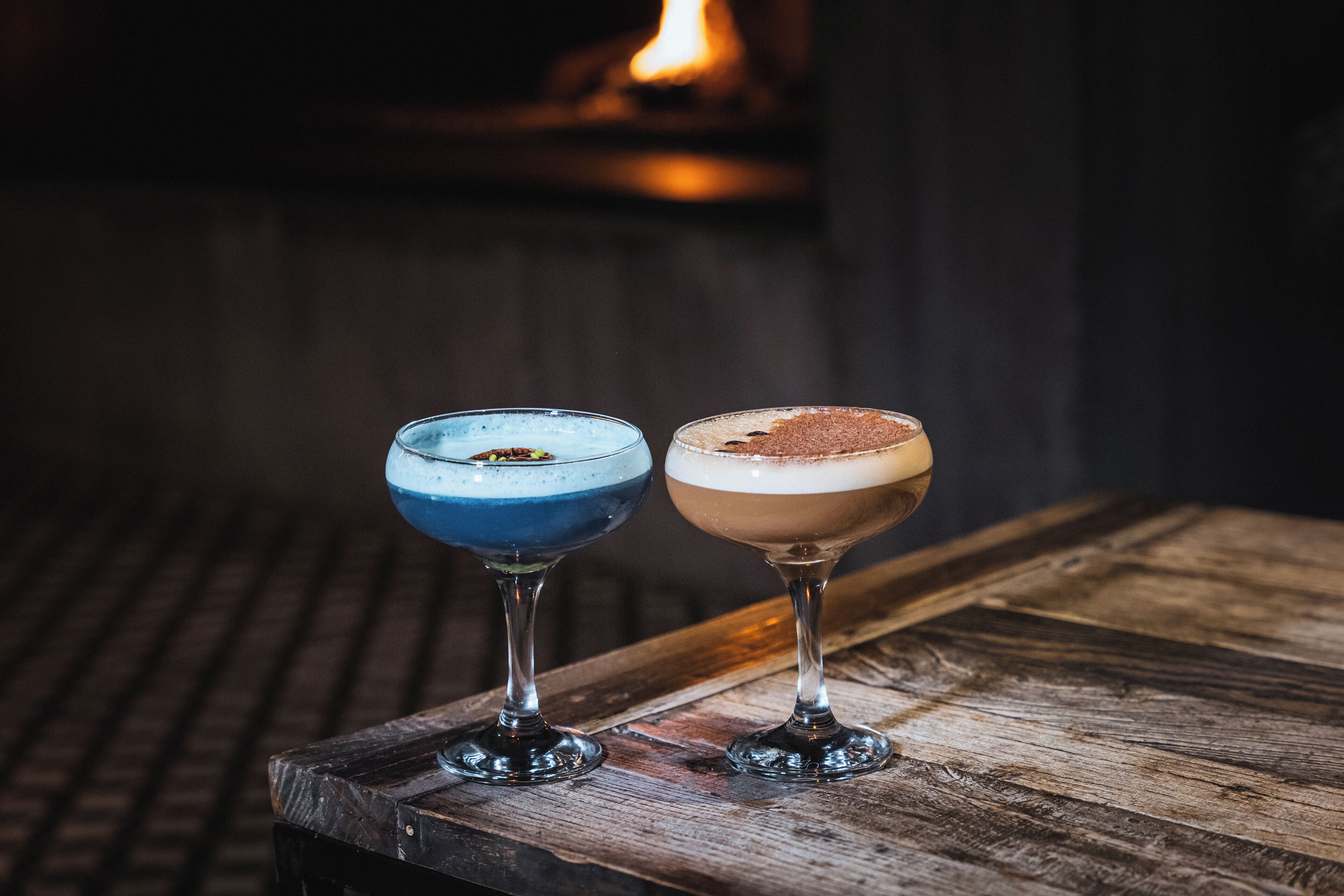 Dos cócteles sobre una mesa de madera con una chimenea al fondo.