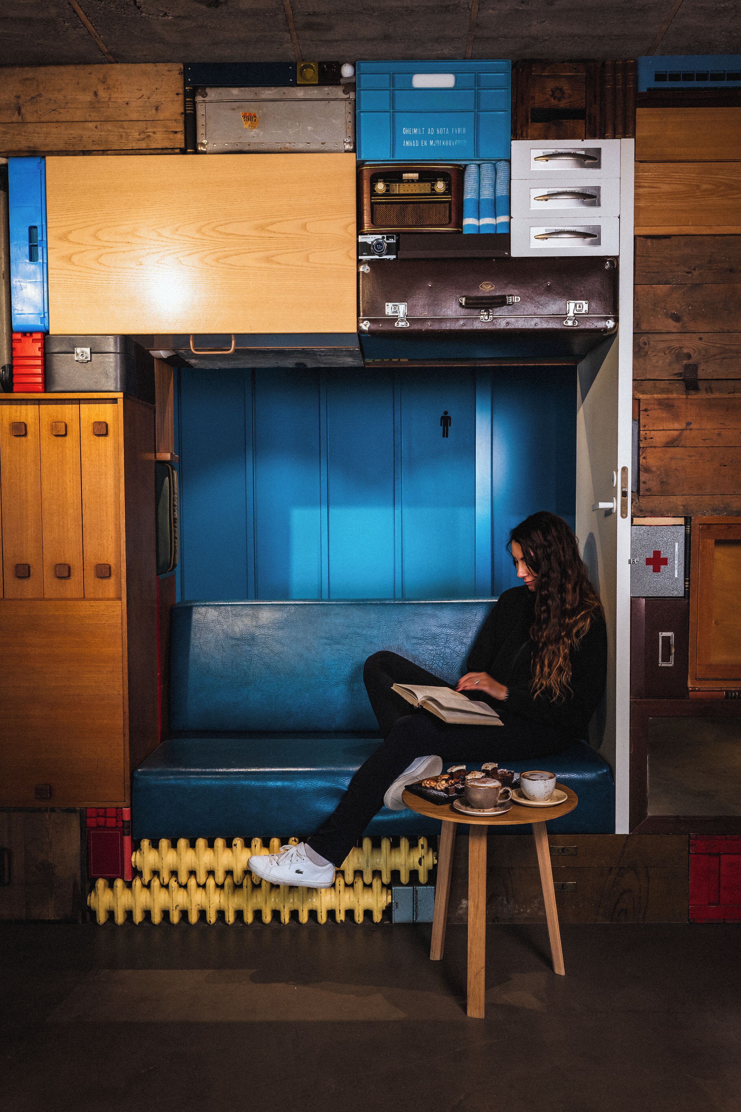 A person sitting on a blue bench reading a book in a room with wooden walls, a wooden cabinet, and a small table with a cup and snacks.