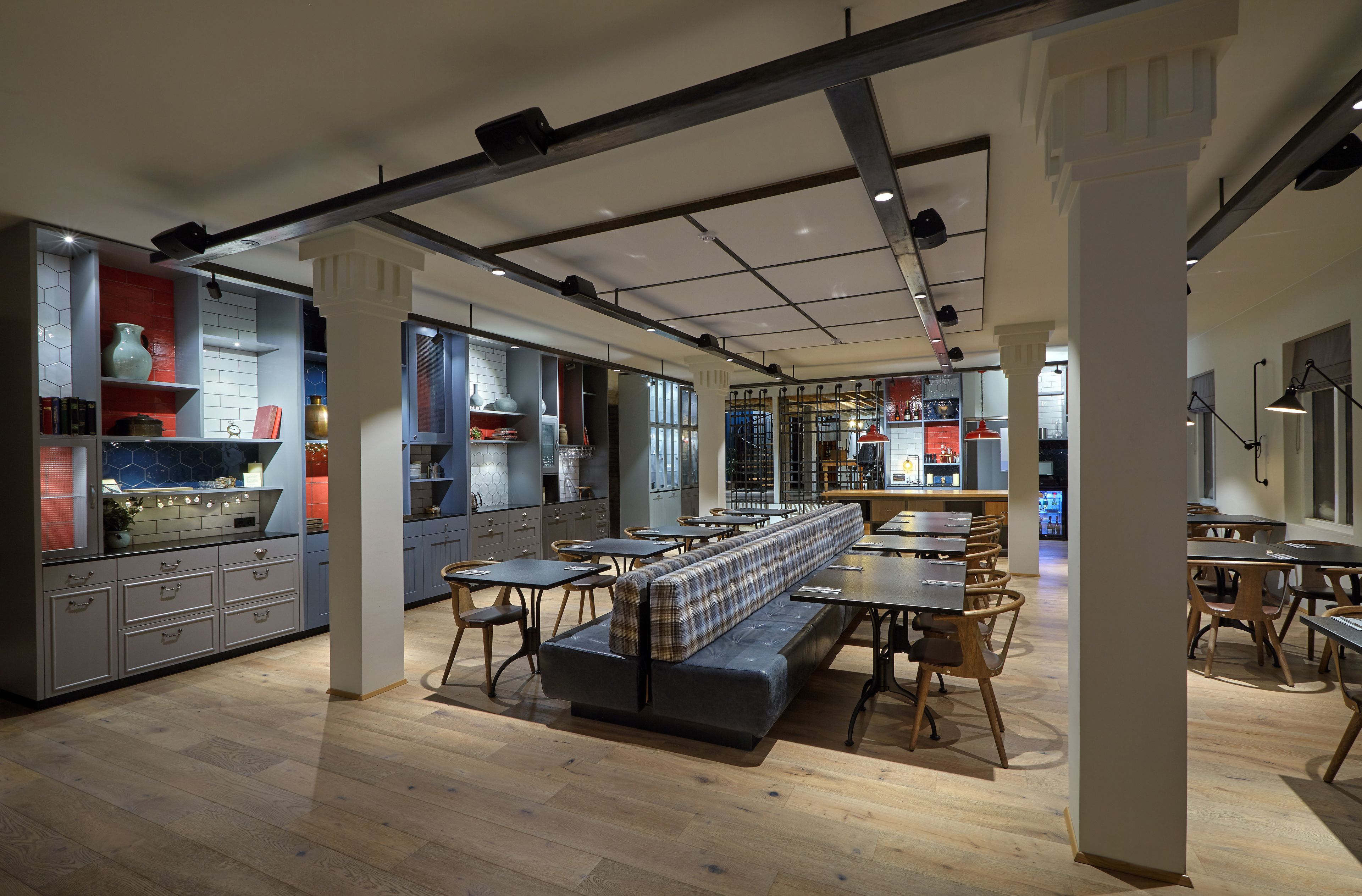 A modern restaurant with a long bench seating area, wooden tables, and a kitchen visible in the background.