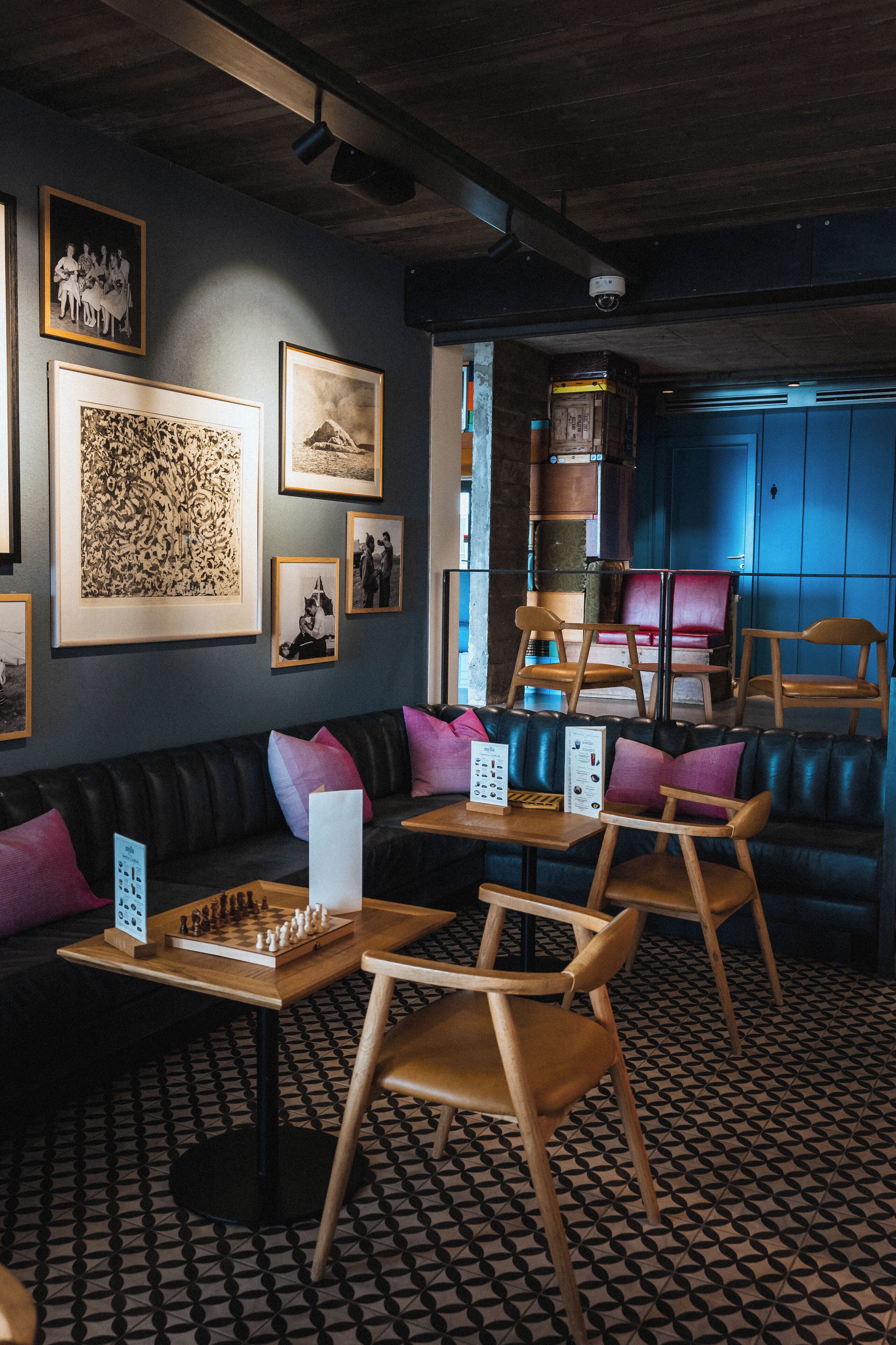 A cozy lounge area with a patterned floor, a black leather sofa, wooden chairs, and framed pictures on the walls.