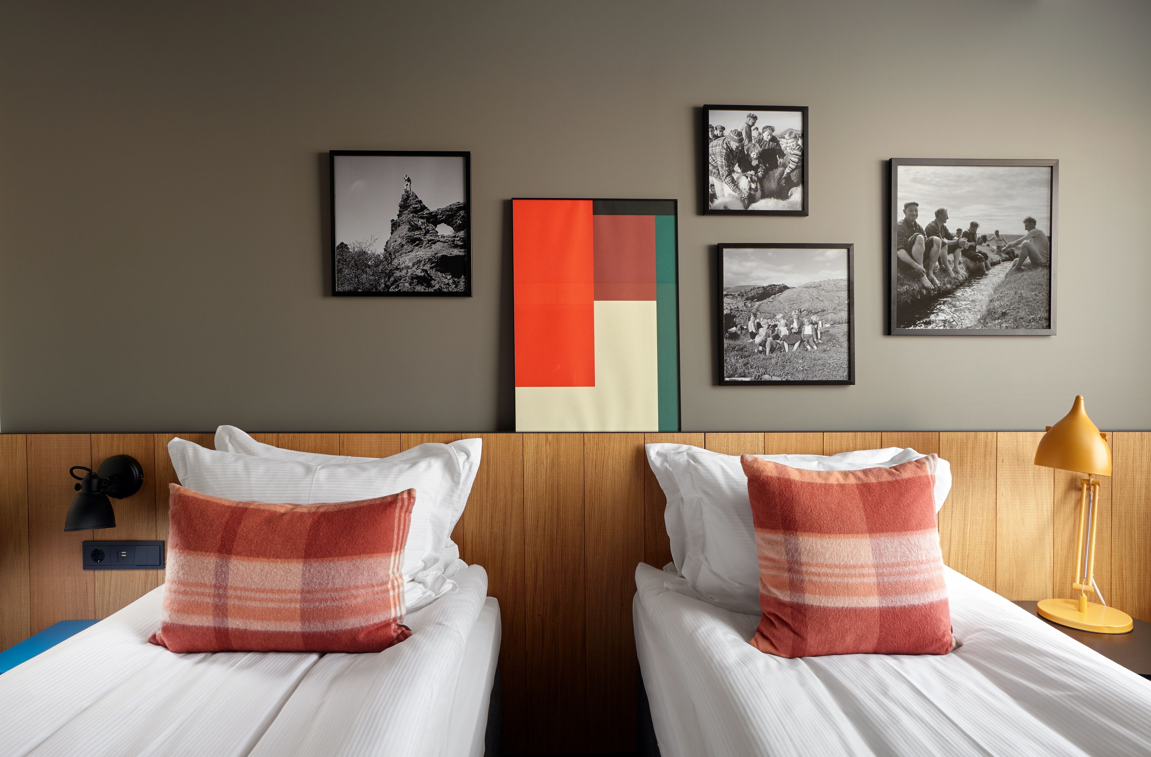 Habitación de hotel con dos camas, un cabecero de madera, una mesita de noche con lámpara y cuatro cuadros enmarcados en la pared.