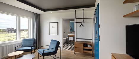 A modern hotel room with a large window, two blue armchairs, a wooden coffee table, a TV, and a blue wardrobe.