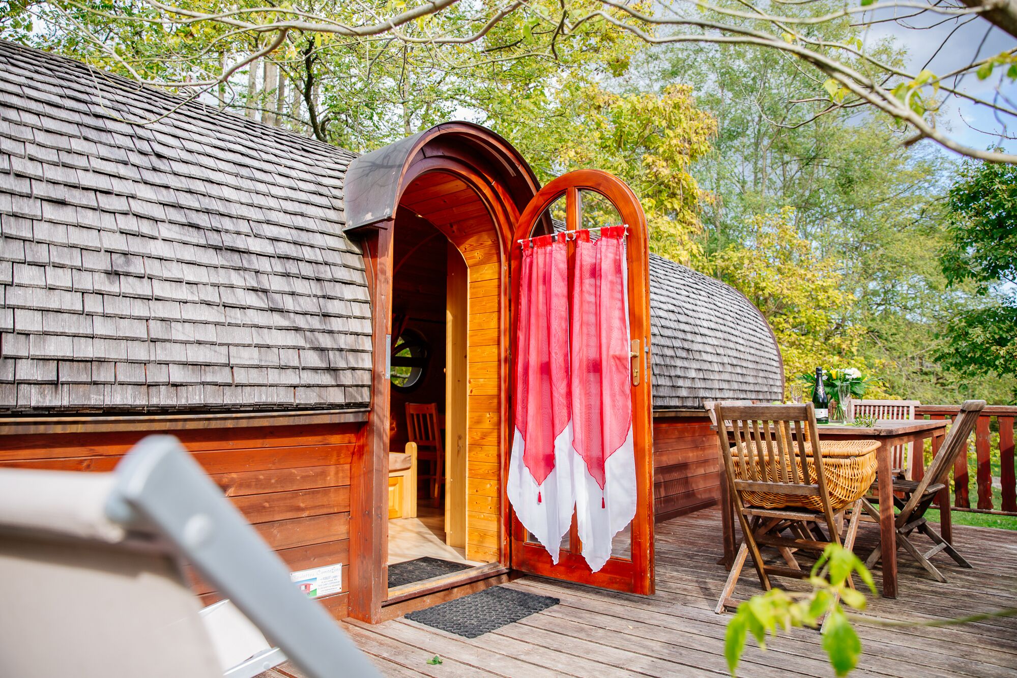 A wooden cabin with a red door, a wooden deck, and a table with chairs.