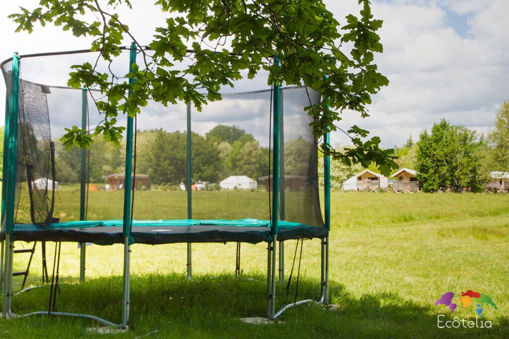 Un trampolín con red de seguridad, rodeado de un campo cubierto de césped, con árboles y casas al fondo.