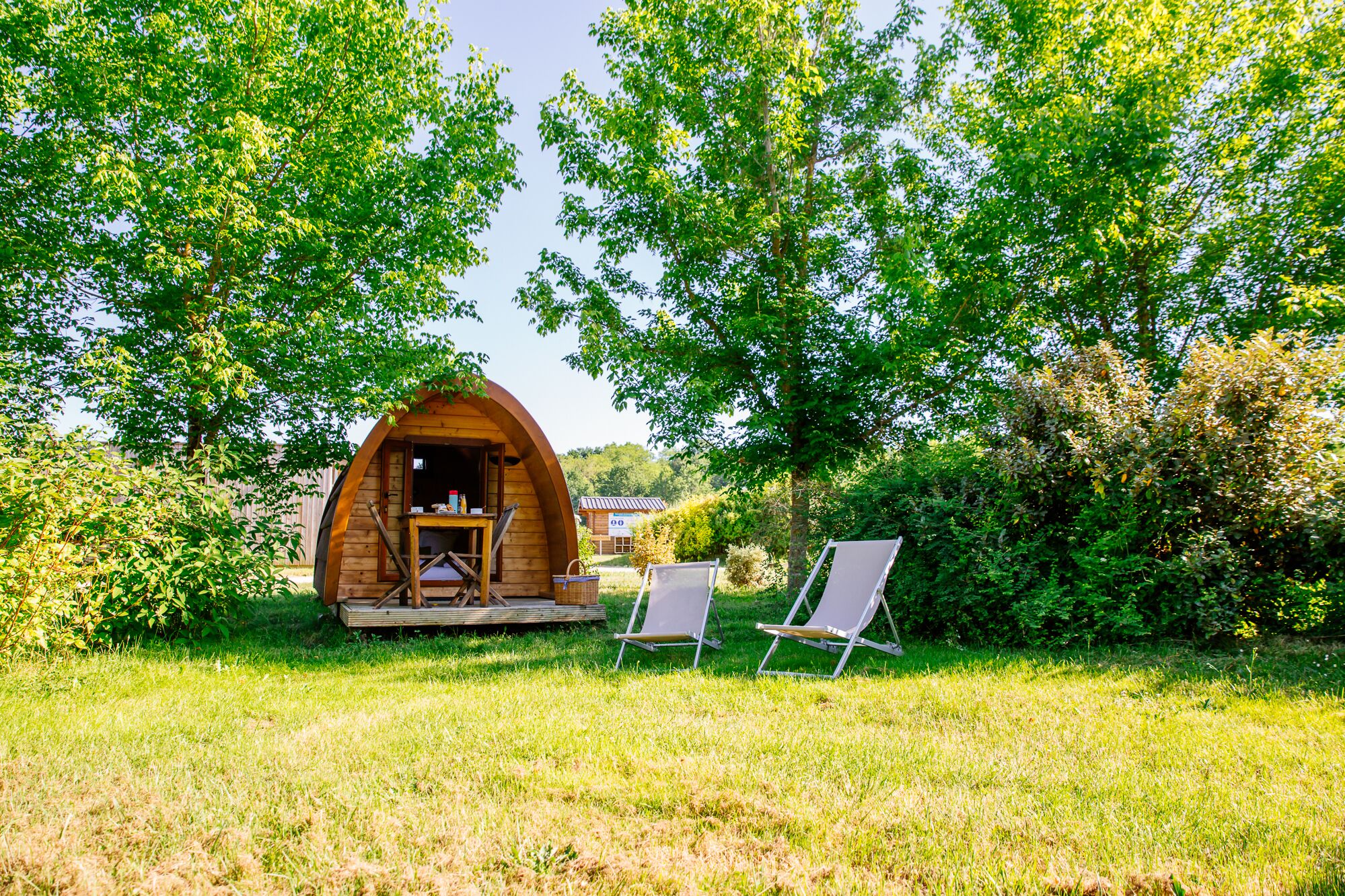 A wooden cabin with a porch, two chairs, and a table in a grassy area with trees.