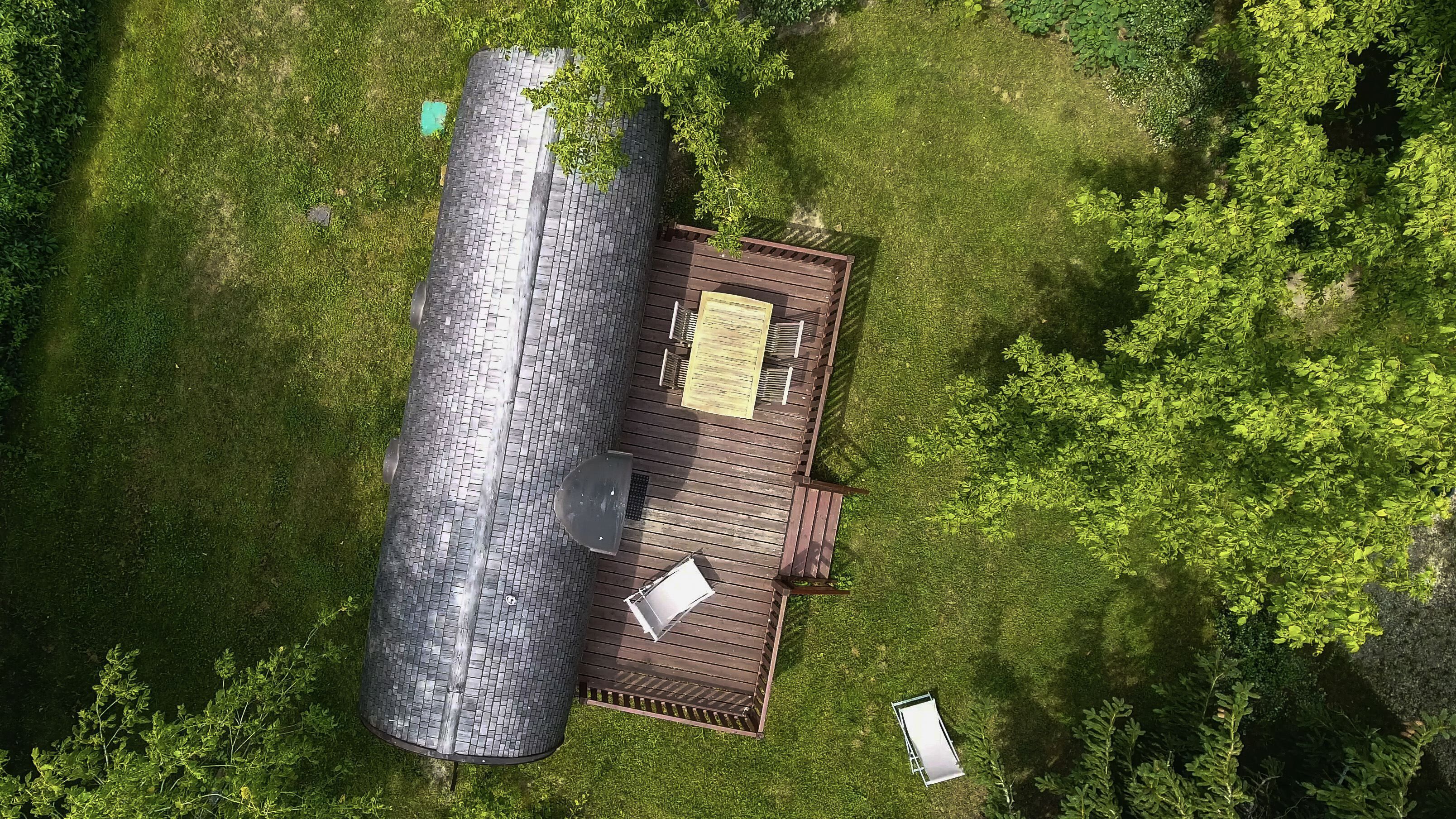 Vue aérienne d’une propriété comprenant une grande structure cylindrique, une terrasse en bois avec une table et des chaises, et une végétation luxuriante tout autour.