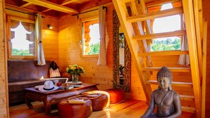 A wooden interior with a meditation statue, a sofa, a table with books and a vase of flowers, and a window with curtains. - Domaine Ecôtelia (Le Nizan)