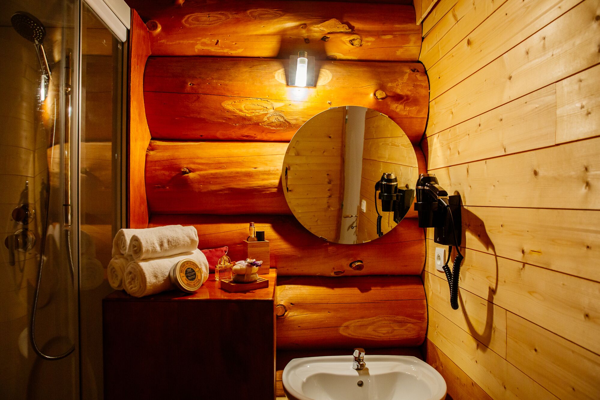 Baño de cabaña de madera con ducha de vidrio, un espejo redondo, un lavabo y un tocador de madera.