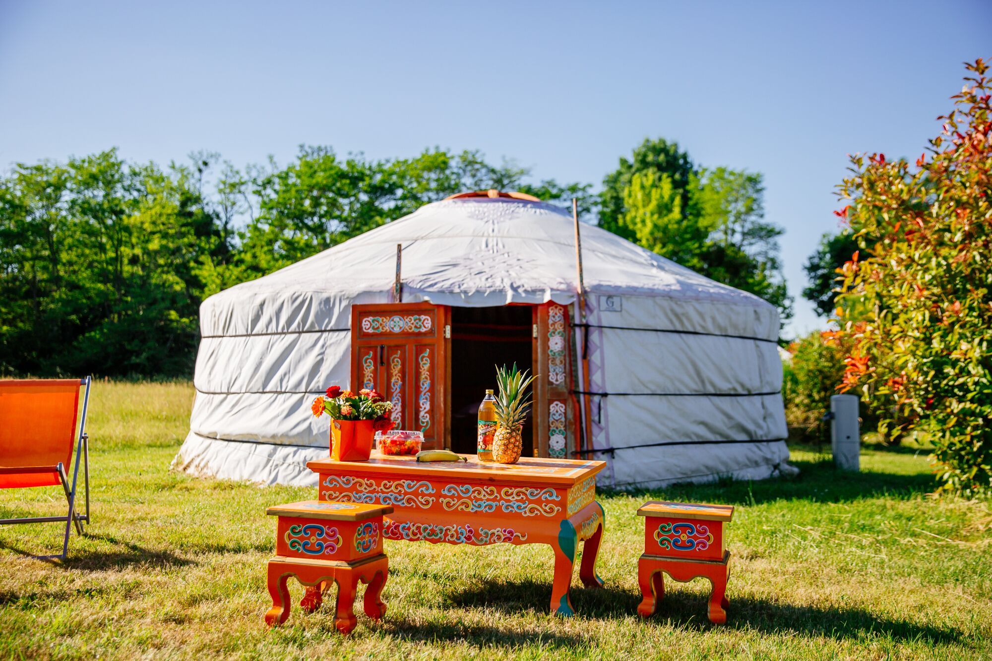 Een joert met een tafel en twee krukken ervoor, omringd door gras en bomen.