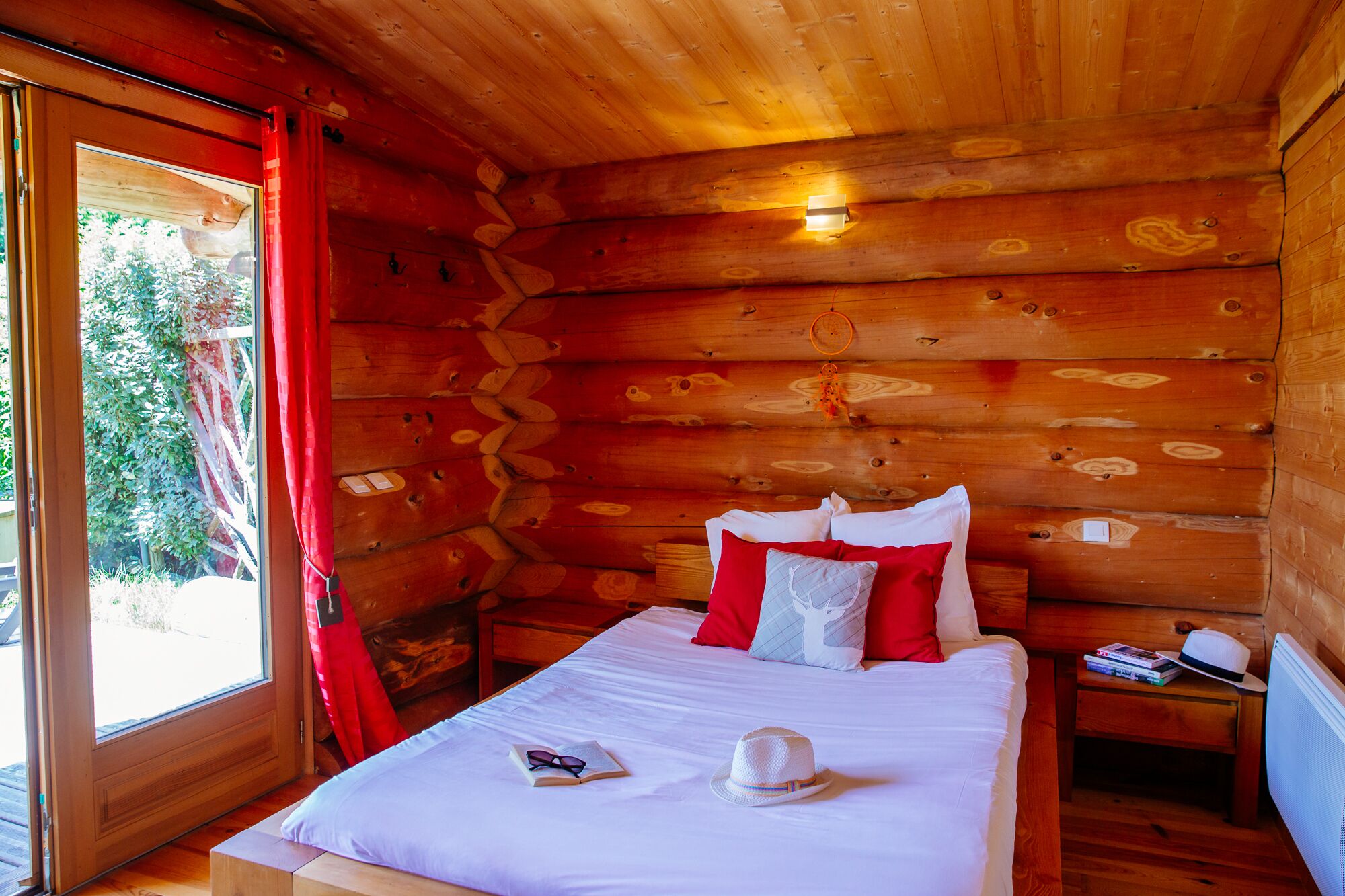 Ein Zimmer in einer Holzhütte mit einem Bett, roten Vorhängen und einem Fenster mit Blick auf Grünflächen.