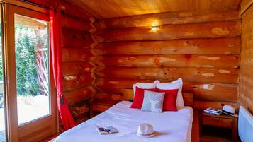 A wooden cabin room with a bed, red curtains, and a window with a view of greenery.