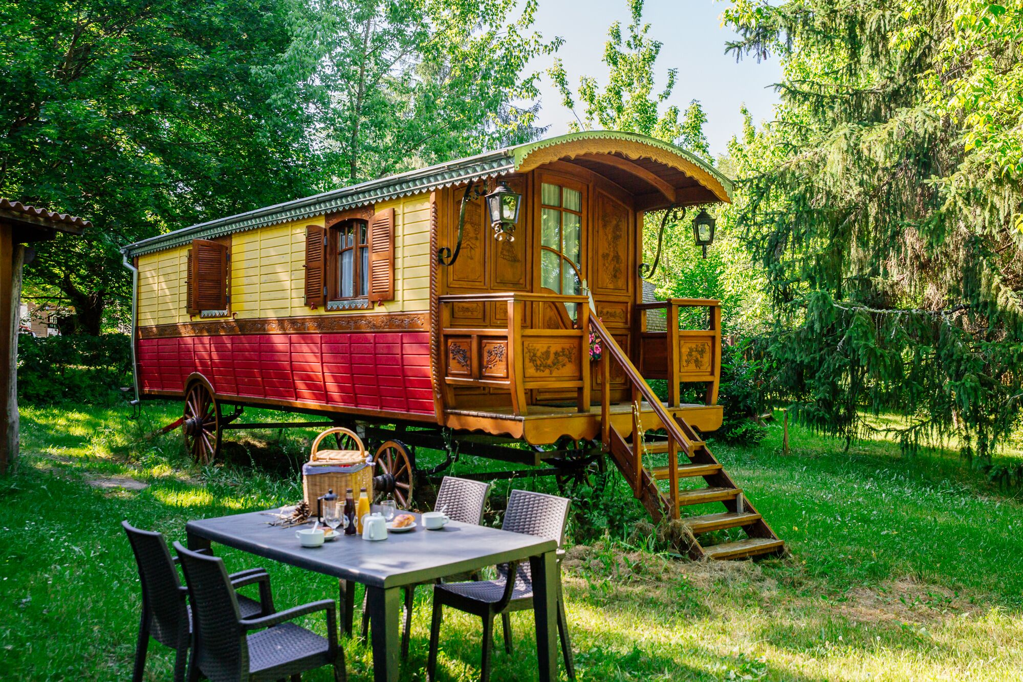 Una casa flotante de madera con exterior rojo y amarillo, una escalera que lleva a una cubierta, y una mesa con sillas dispuestas al frente.