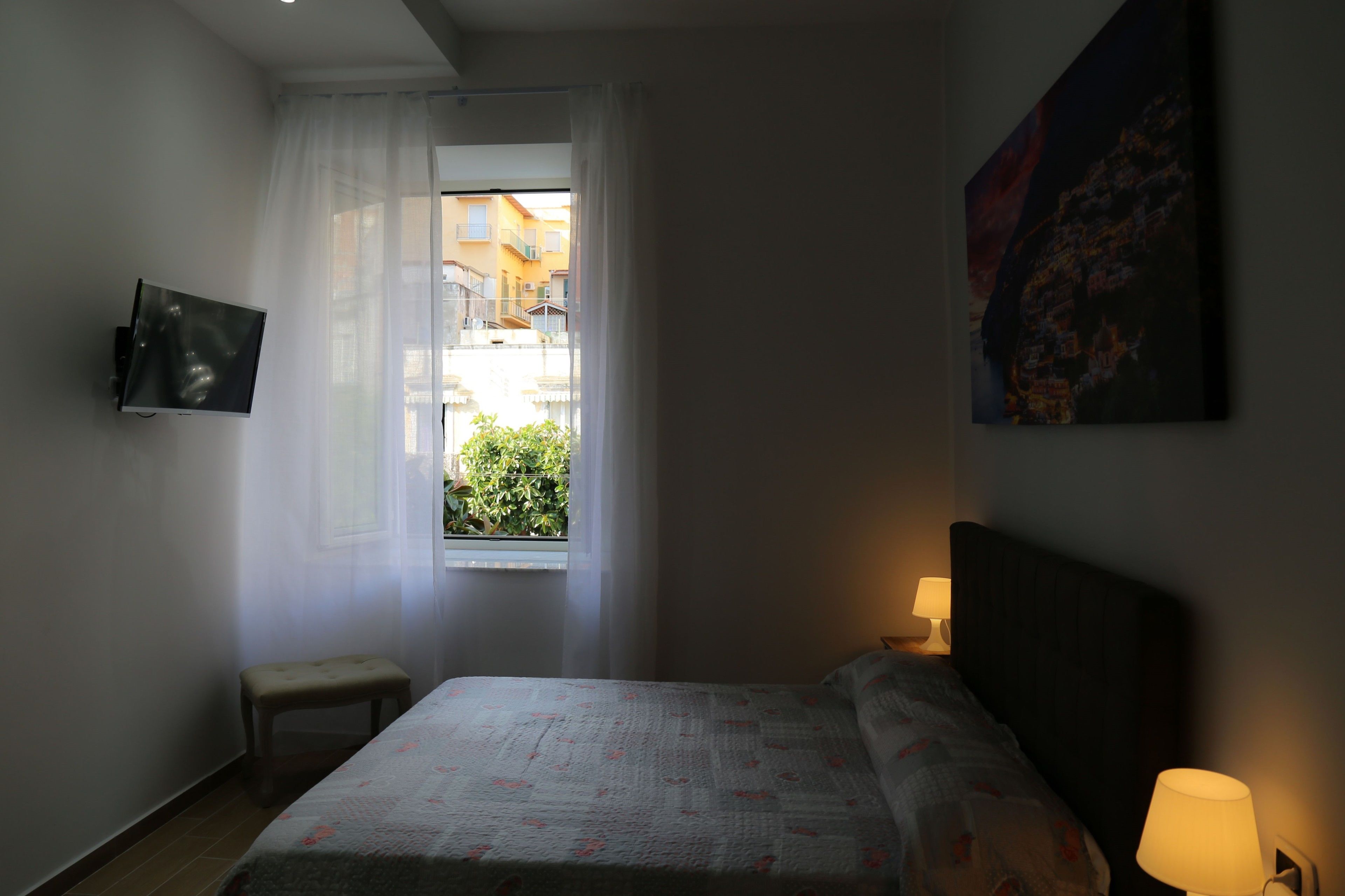 A bedroom with a bed, a small table, a TV mounted on the wall, a window with sheer curtains, and a nightstand with a lamp.