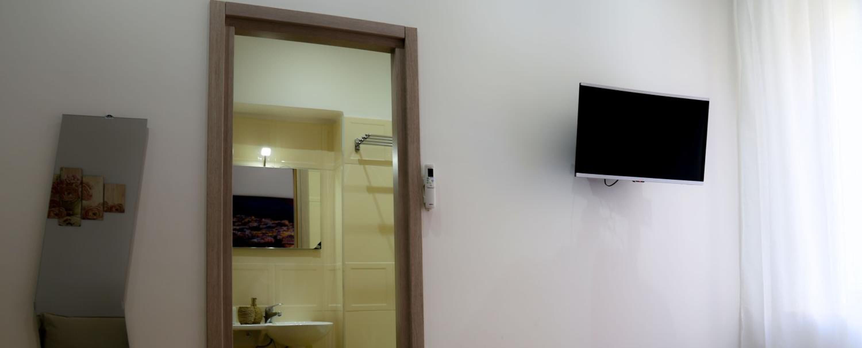 A room with a white wall, a mounted TV, a door leading to a bathroom, and a small shelf with a vase.