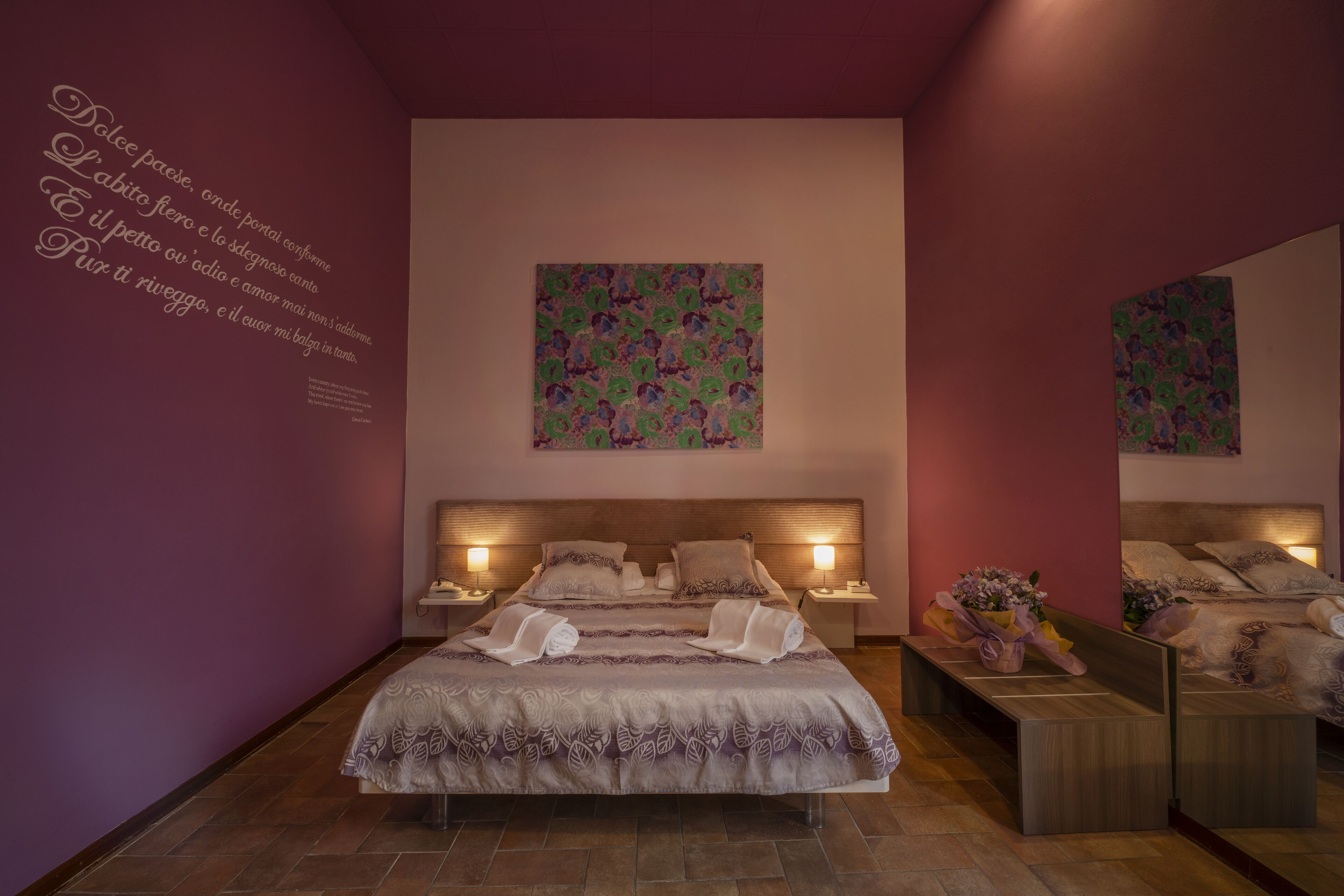 Une chambre à coucher avec un grand lit, deux tables de chevet, un miroir et un mur décoratif orné d’un texte et d’une peinture florale.