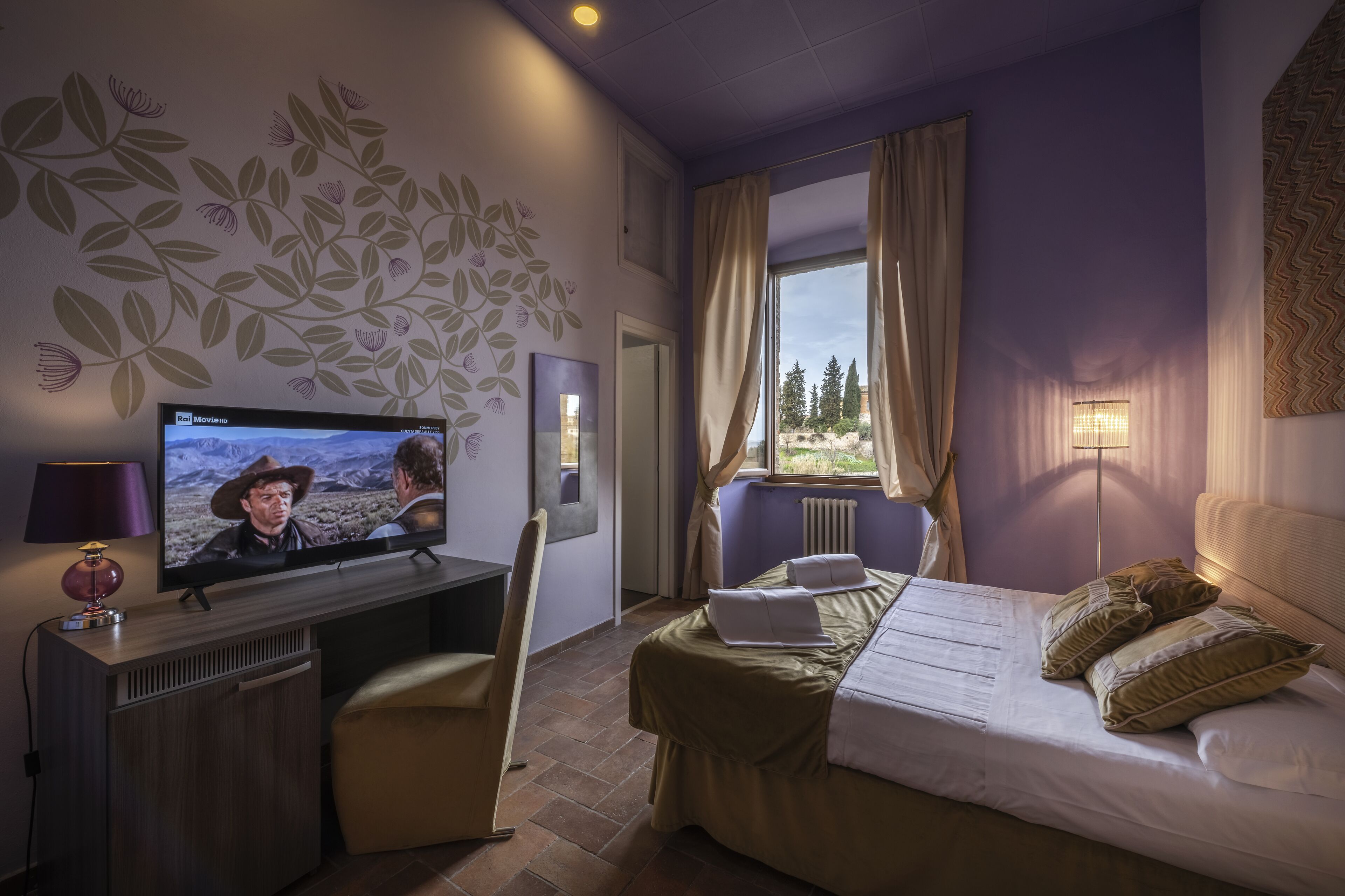 Habitación de hotel con cama, escritorio con televisor, una silla y ventana con cortinas.