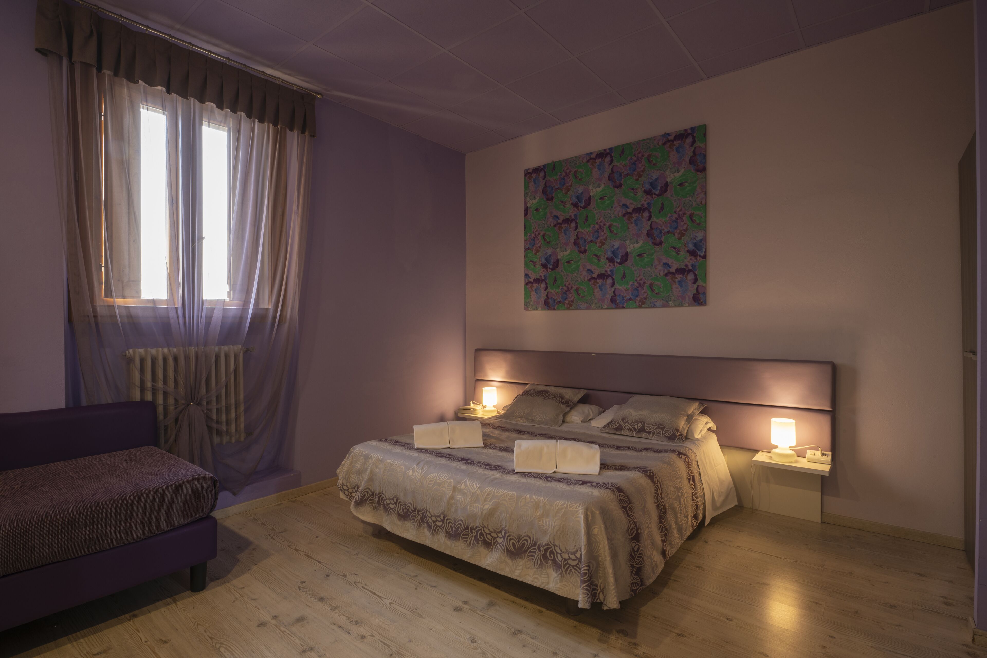 A bedroom with a large bed, two bedside tables with lamps, a window with curtains, a radiator, and a colorful abstract painting on the wall.