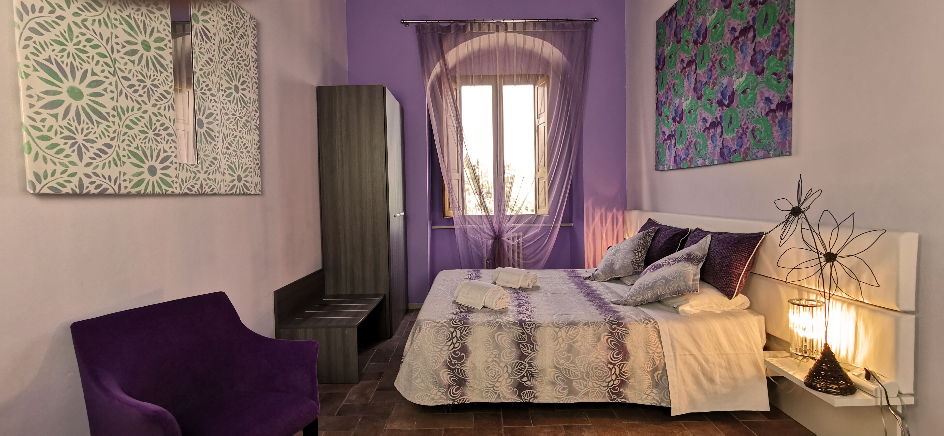 Een slaapkamer met een bed, een paarse stoel, een kledingkast, een raam met gordijnen en een kunstwerk aan de muur.
