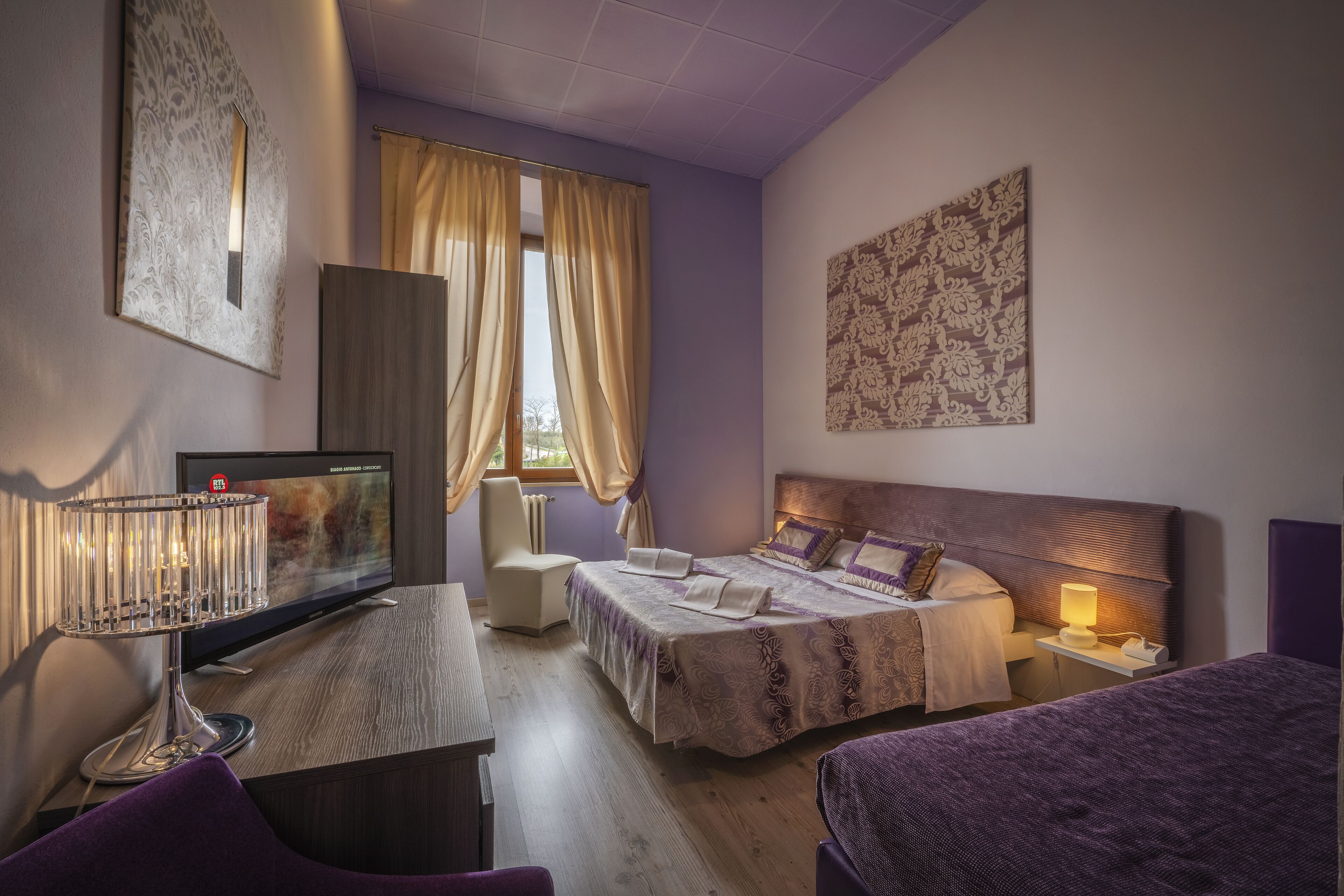 Habitación de hotel con cama, escritorio, silla, televisor y ventana con cortinas.
