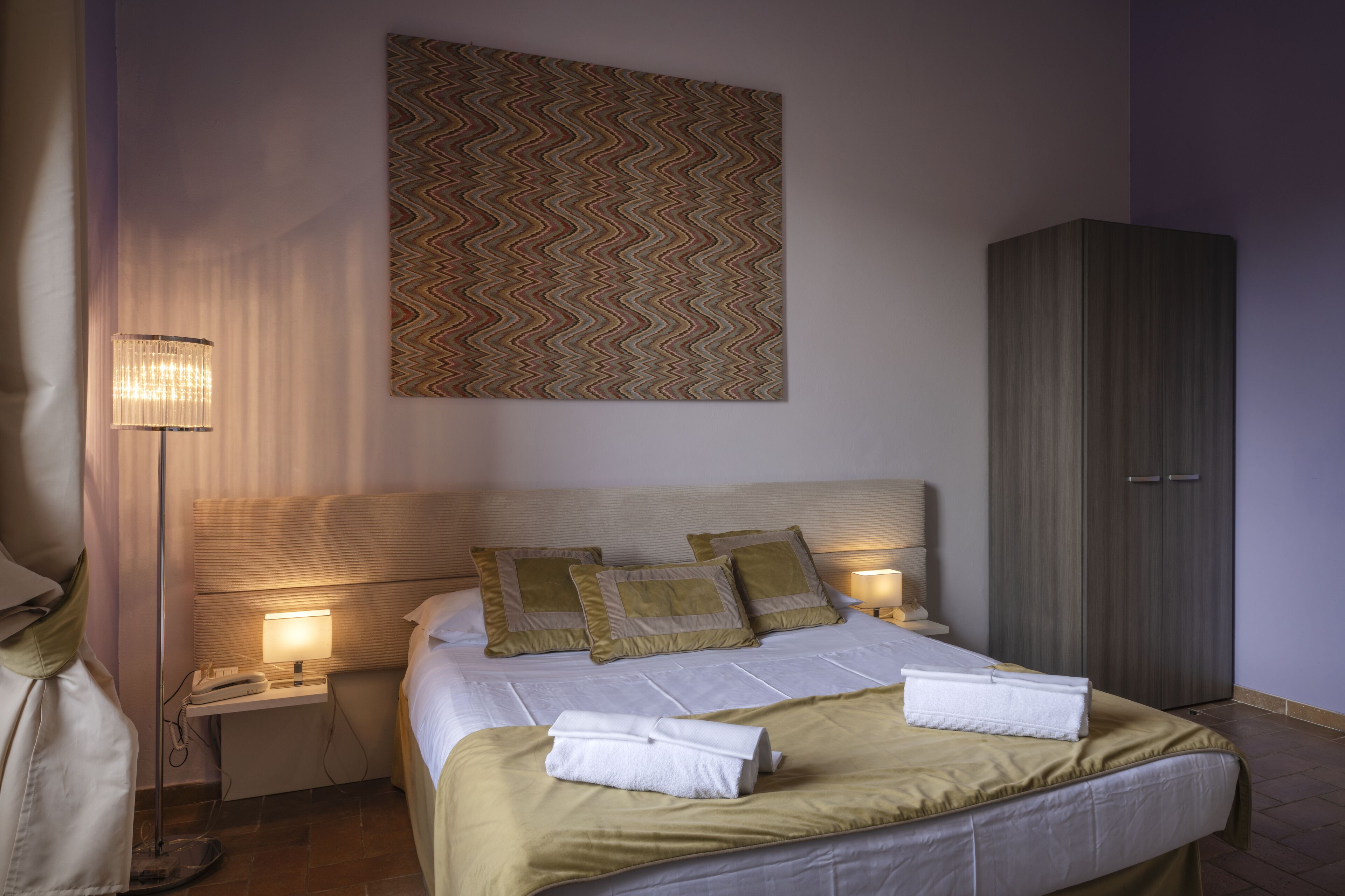 Una cama bien tendida con una colcha verde y dos almohadas, una lámpara de noche, una obra de arte en la pared y un armario.