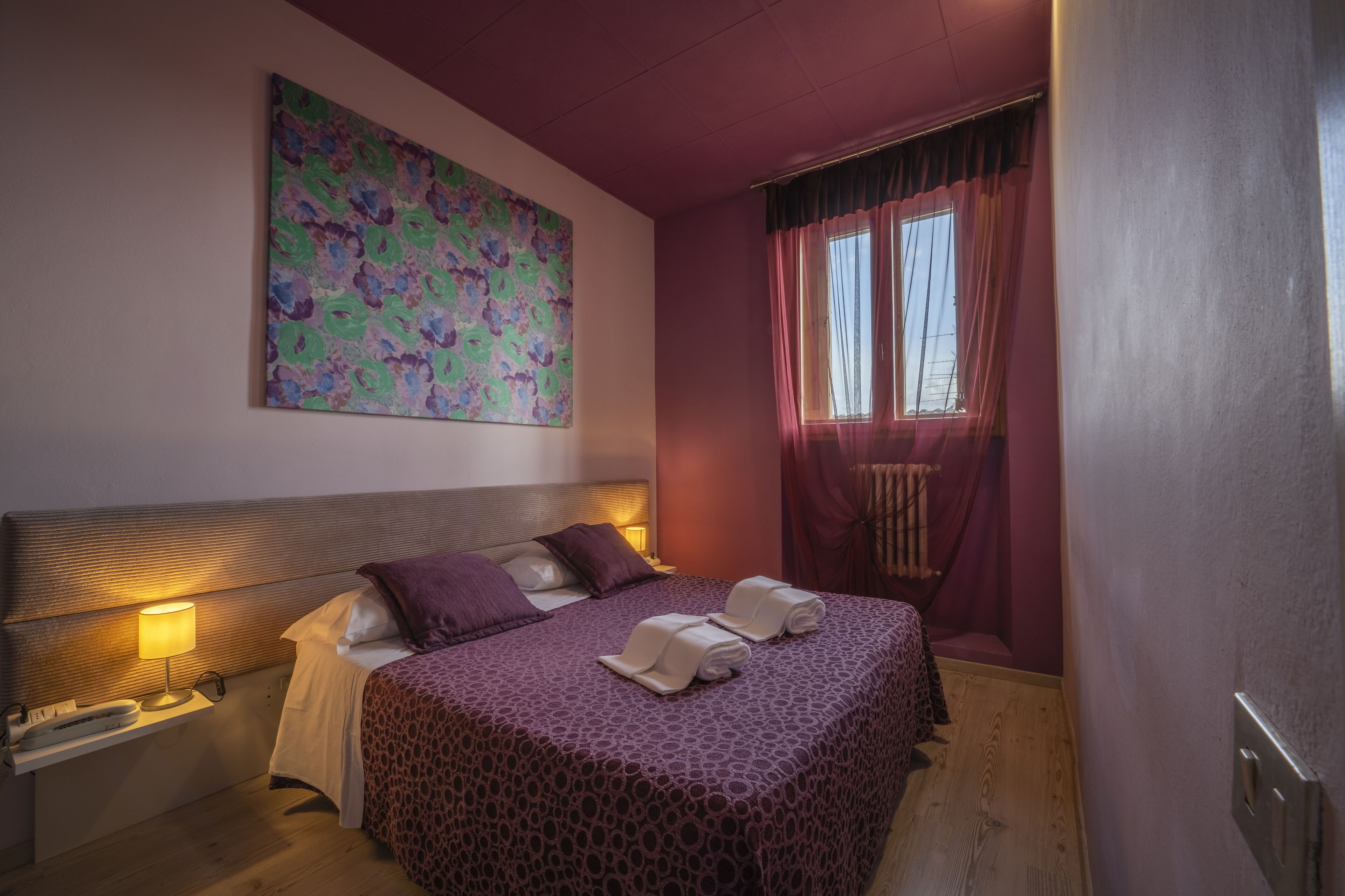 Ein Schlafzimmer mit einem großen Bett, einem Kopfteil, einem Nachttisch mit Lampe, einem Fenster mit Vorhängen und einem farbenfrohen, abstrakten Gemälde an der Wand.