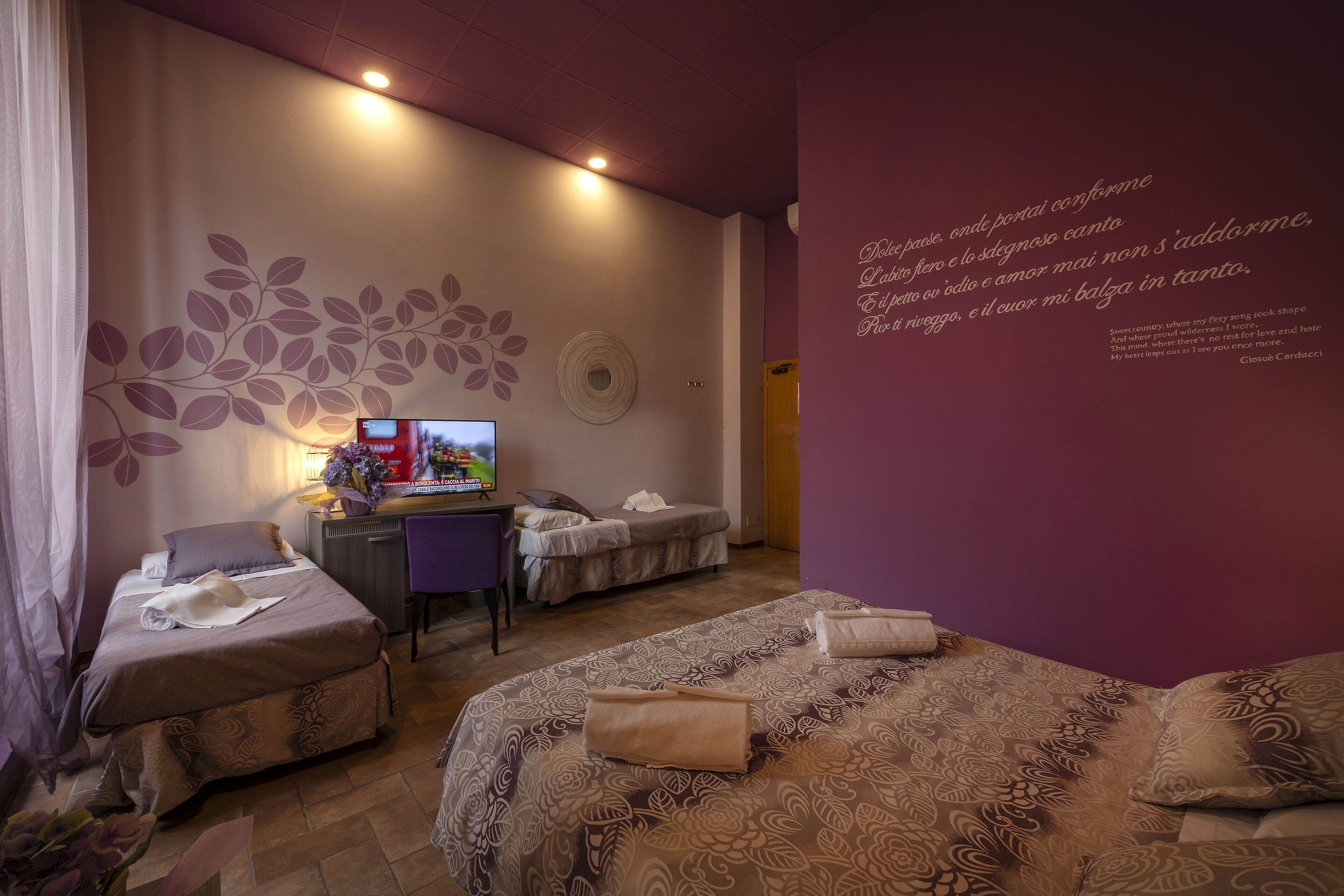 A hotel room with two beds, a desk, a TV, and a wall with decorative text.