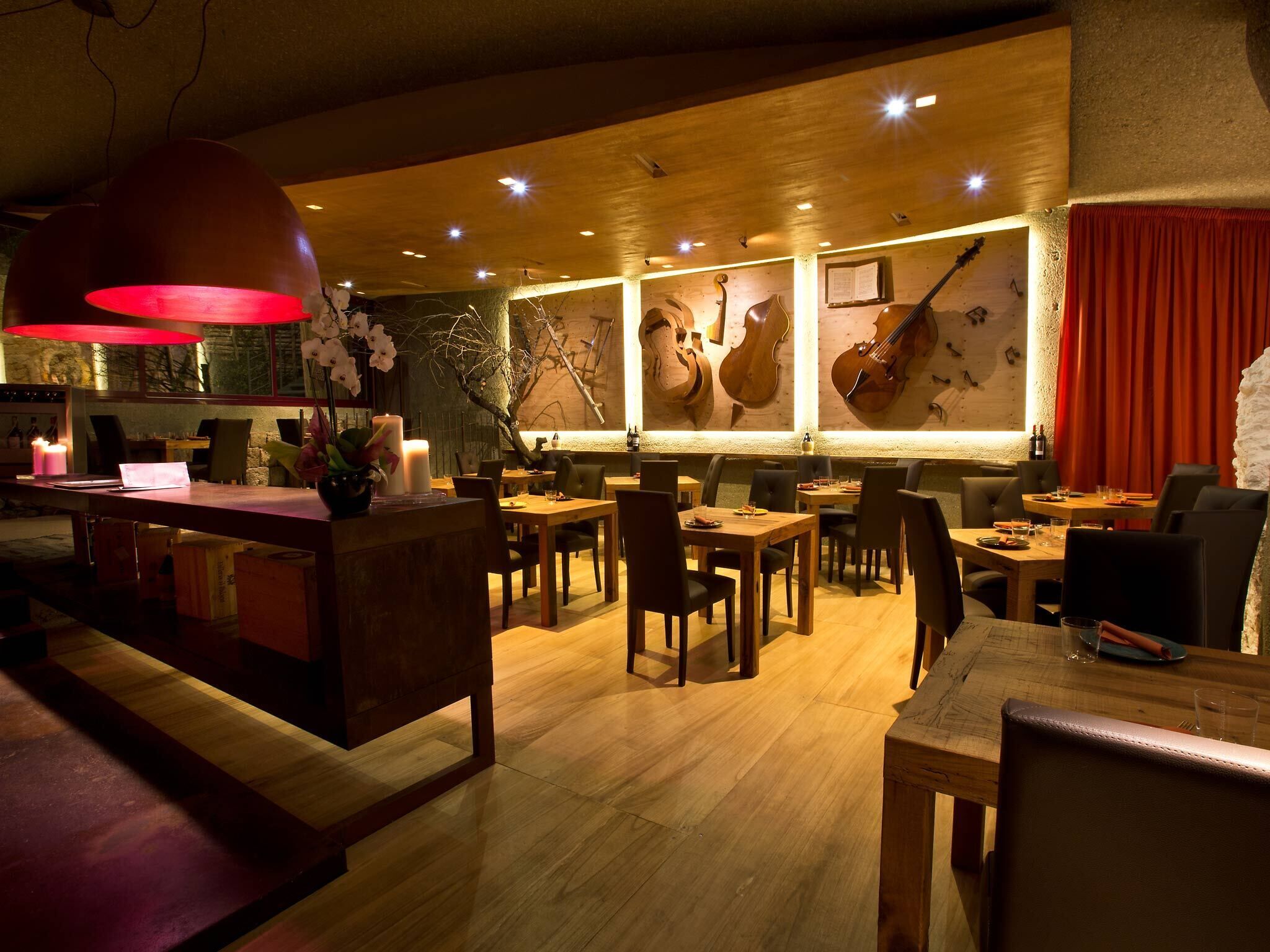 A restaurant interior with wooden tables and chairs, a bar area with a red pendant light, and wall-mounted musical instruments.