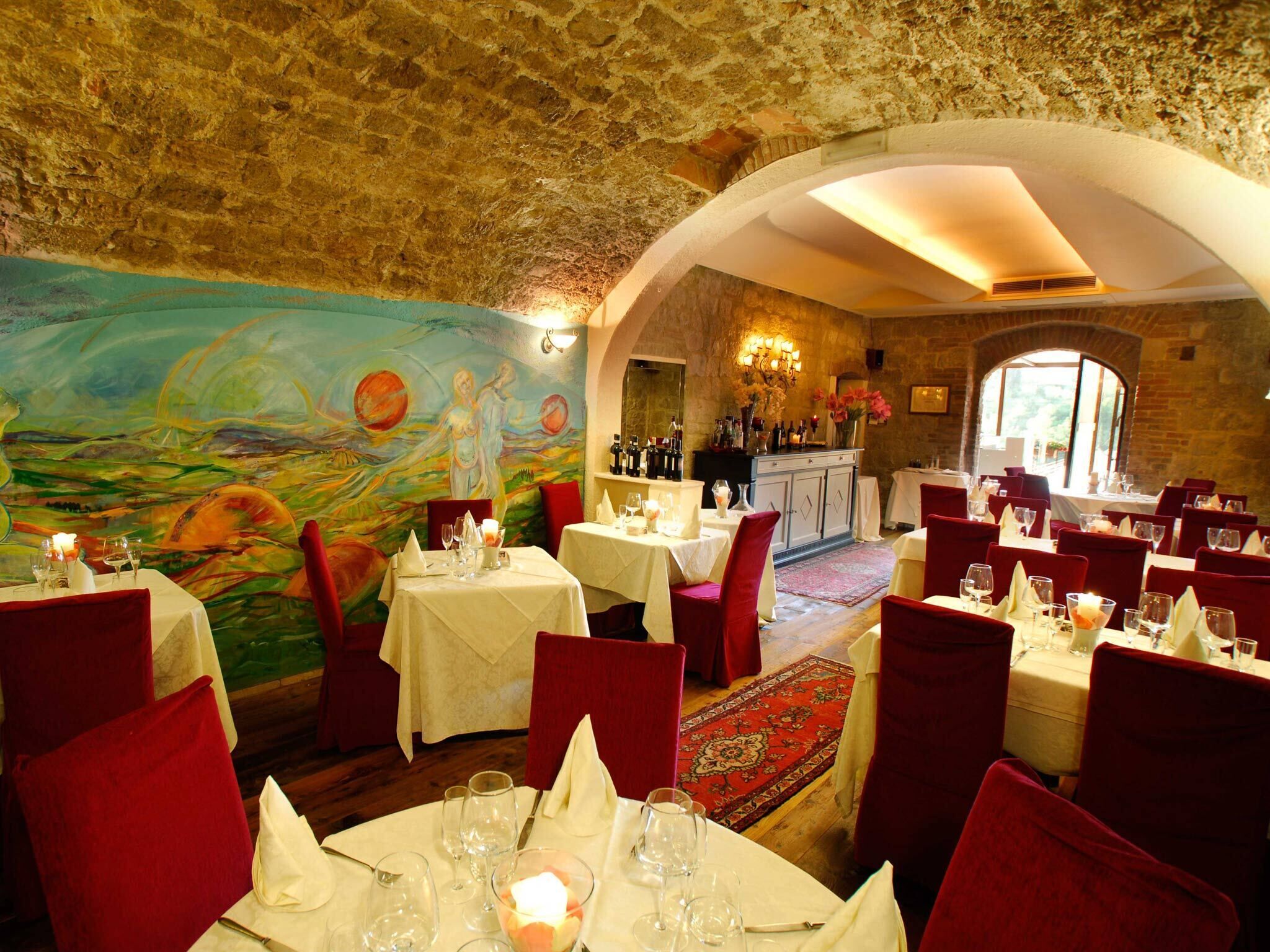 Een restaurant met stenen muren, rode fluweelstoelen en een muurschildering. Tafels zijn gedekt met witte tafellinnen en kaarsen.